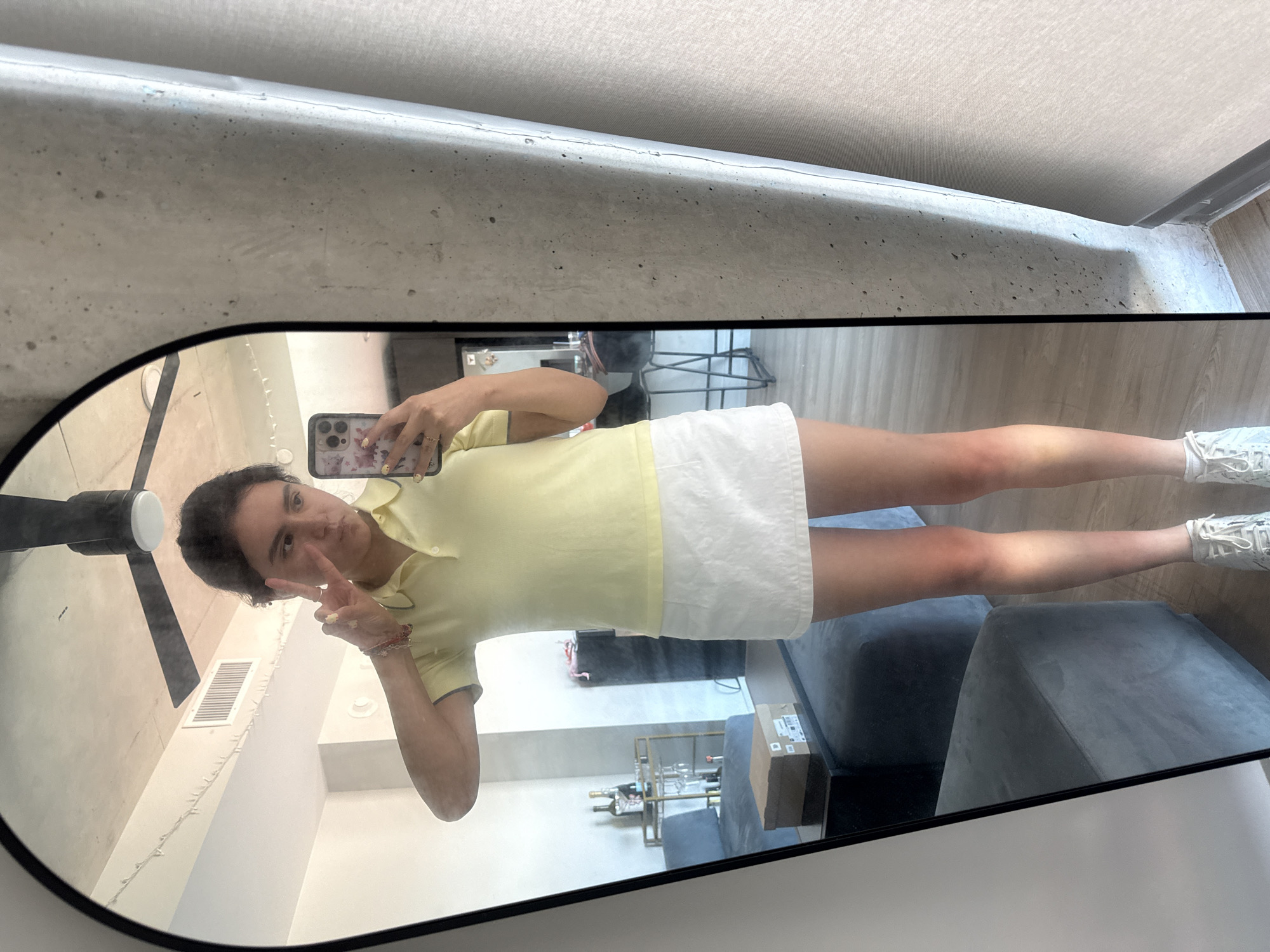A selfie taken in a mirror reveals a woman in a yellow collared shirt and white shorts. She holds a phone in her right hand, its back adorned with a patterned case. Her left hand displays a peace sign near her face. The setting is a modern interior space with a ceiling fan visible in the upper-left corner. The reflection shows a view into a living area with minimalist furniture, including a gray ottoman and a metal wine rack holding bottles and glasses. Given the contemporary aesthetic and visible elements, it is reasonable to infer that the location is within a modern apartment or residence in Austin, United States of America.