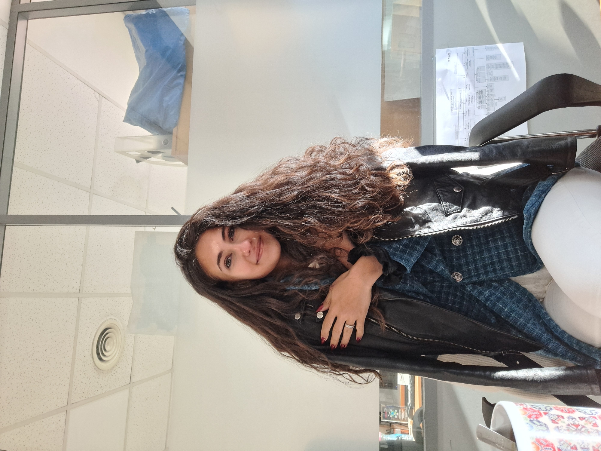 A young woman sits indoors, possibly in an office setting. She is facing the camera with a slight smile. The woman has long, curly dark brown hair and wears a black leather jacket over a blue patterned shirt. The jacket is resting on a chair with a hanger, and she appears to be sitting in the chair. She has manicured nails with dark red polish and wears a ring on one of her fingers. The background features a paneled ceiling with air vents and a wall with a window. Through the window, part of a building can be seen. A document hangs on the wall. There is not enough information to determine the city of Sofia, Bulgaria.