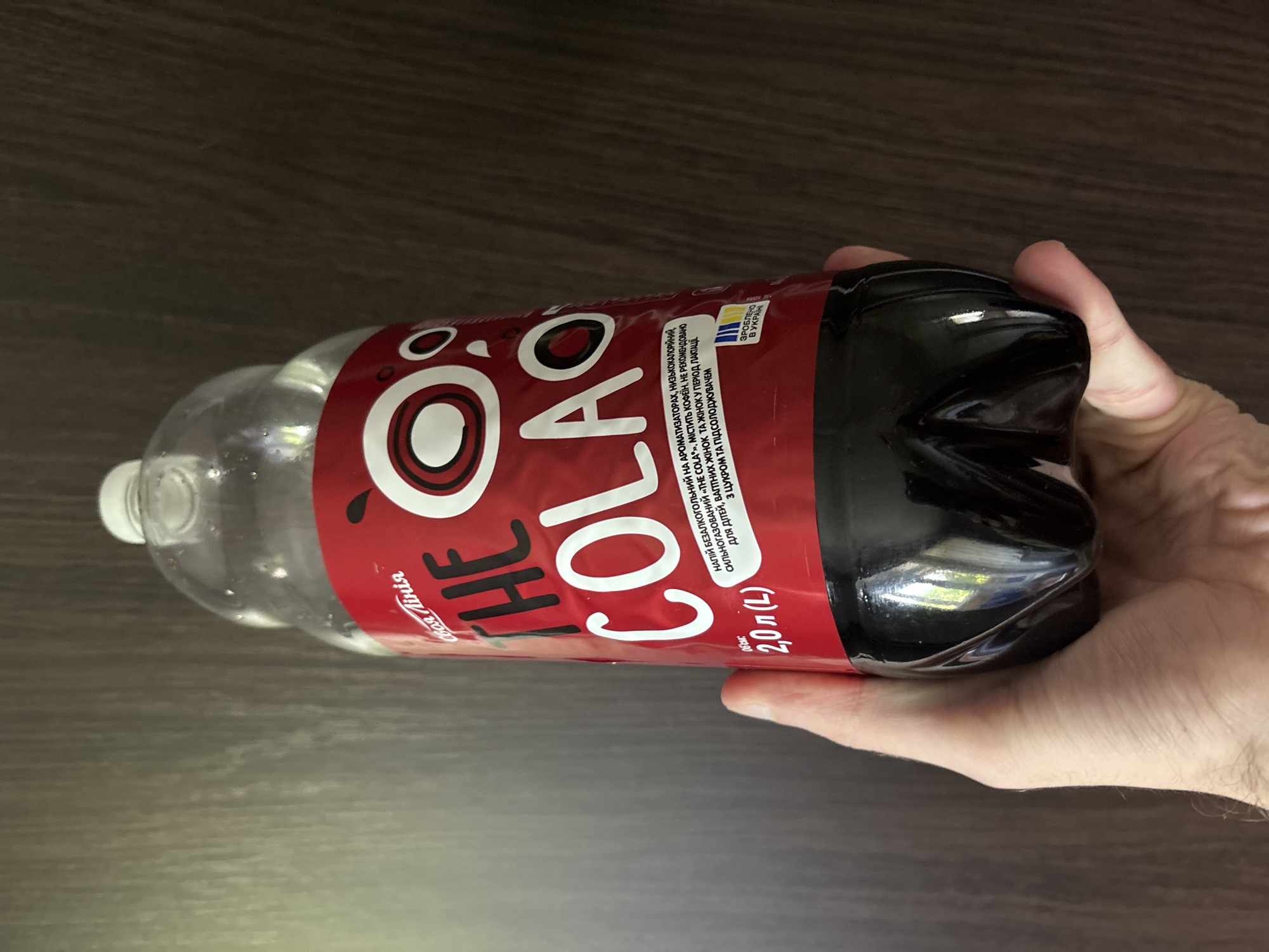 A hand is holding a large, 2-liter bottle of a dark-colored beverage, likely soda, with a red and white label. The bottle is lying on its side on a dark wooden surface. The label prominently displays the word "COLAO" in large, stylized white letters, with "THE" written smaller above it. There is a graphic of a red circle with a white outline and a white inner circle on the label.
On the label, there is text in Ukrainian. One section reads "НАПІЙ БЕЗАЛКОГОЛЬНИЙ НА АРОМАТИЗАТОРАХ, НИЗЬКОКАЛОРІЙНИЙ. СИЛЬНОГАЗОВАНИЙ «THE COLA», МІСТИТЬ КОФЕЇН, НЕ РЕКОМЕНДОВАНО ДЛЯ ДІТЕЙ, ВАПТНИХ ЖІНОК ТА ЖІНОК У ПЕРІОД ЛАКТАЦІЇ З ЦУКРОМ ТА ПІДСОЛОДЖУВАЧЕМ". This translates to "NON-ALCOHOLIC DRINK WITH FLAVORINGS, LOW-CALORIE. HIGHLY CARBONATED 'THE COLA', CONTAINS CAFFEINE, NOT RECOMMENDED FOR CHILDREN, PREGNANT WOMEN AND WOMEN DURING LACTATION WITH SUGAR AND SWEETENER".
Another part of the label indicates "2,0 л (L)" for the volume and has a blue and yellow Ukrainian flag icon with the text "ЗРОБЛЕНО В УКРАЇНІ", meaning "MADE IN UKRAINE". The bottle has a white cap. The lighting suggests an indoor setting, possibly with artificial light, and the overall impression is a casual, everyday scene.