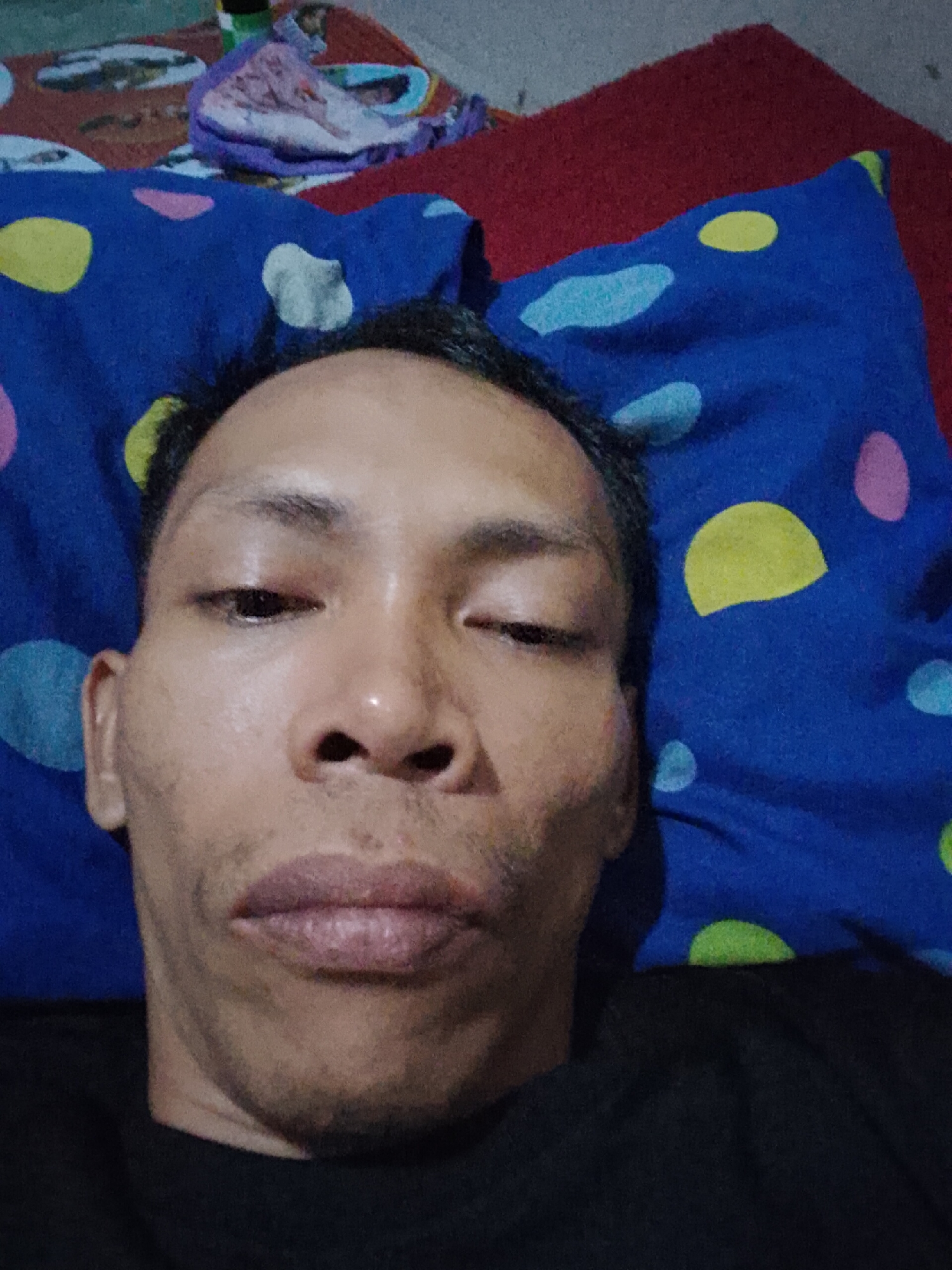 The image shows a close-up, frontal view of a person lying down, with their face taking up most of the frame. The person appears to be indoors, likely in a bedroom. They are lying on a blue pillow with colorful polka dots, and a red textured surface, possibly a blanket or bedspread, is visible behind the pillow. A small pile of colorful fabric, possibly clothing or laundry, is also present in the upper portion of the background. The lighting appears to be artificial and the image quality is somewhat grainy, suggesting it was taken with a mobile phone camera in low light conditions. The person's facial features are distinct, with dark hair, prominent eyebrows, and visible skin texture. No specific location in Bukit Ketok, Indonesia, can be determined from the visual information provided.