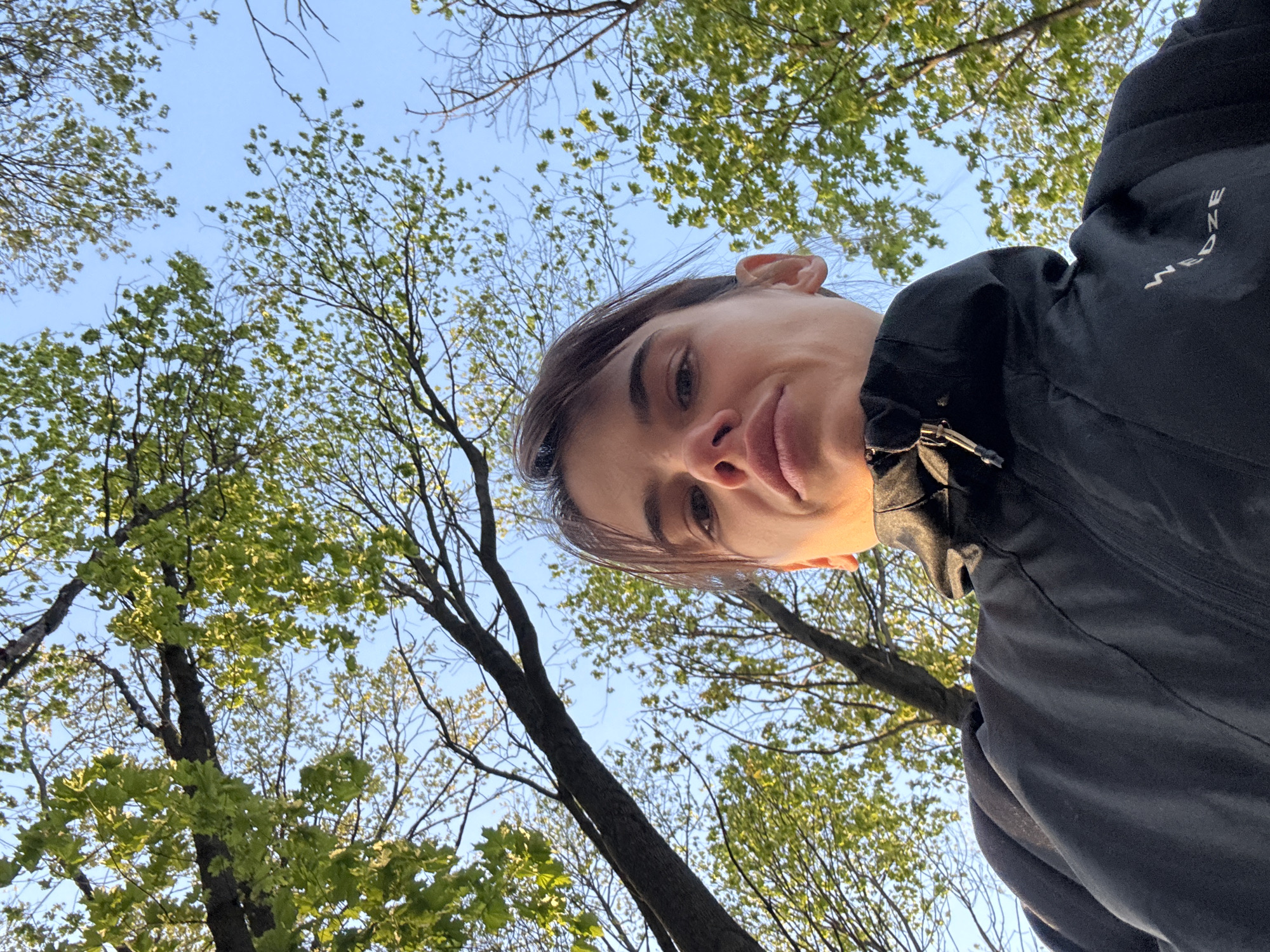An upward-angled, worm's-eye view shot captures a person's upper body and face against a natural backdrop. The individual, appearing to be taking a selfie, looks directly into the camera from the top of the frame. Their face shows a neutral to slightly pouting expression, with dark eyes and full lips. Dark hair frames their face. They are wearing a dark, possibly black, jacket. On the right side of the jacket, near the shoulder/arm, the white text "W E D Z E" is visible vertically.
The background is dominated by a clear, bright blue sky. Numerous trees, with some branches bare and others adorned with light green, newly unfurling leaves, frame the sky, indicating a spring or early summer season. The bright natural sunlight suggests it is daytime. The setting is an outdoor, verdant environment, likely a park or forest, in Rostov-on-Don, Russia. The overall atmosphere is fresh and natural.