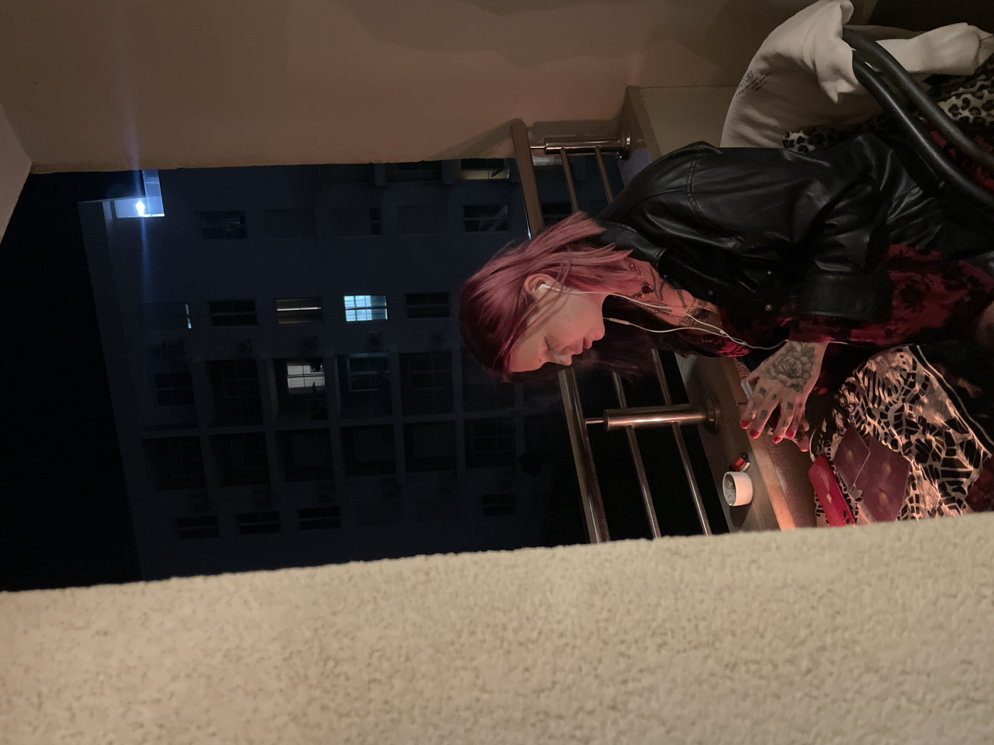 On a residential balcony in Boreti, Montenegro, at night, a female individual with medium-length pink hair is seated, facing left, with her gaze directed downward. She wears a black leather jacket over a dark, possibly red patterned garment, and has visible tattoos on her hands. White wired earbuds are inserted into her ears. A wisp of smoke rises near her face, indicating she is smoking. Her fingernails are painted red.
In front of her, on a patterned cloth, are several dark, patterned cards, resembling tarot cards, next to a red oblong object. A white cup or ashtray and a red lighter rest on a ledge to her left. She is seated in a dark chair, over which a light-colored sweatshirt and a leopard-print fabric are draped. A silver-toned metal balcony railing spans the mid-ground.
The immediate area of the balcony is illuminated by a warm, artificial light source. In the background, under a dark night sky, another multi-story residential building is visible, with several windows illuminated by blue or white interior lights. A light-colored, textured wall or pillar frames the left side of the scene.