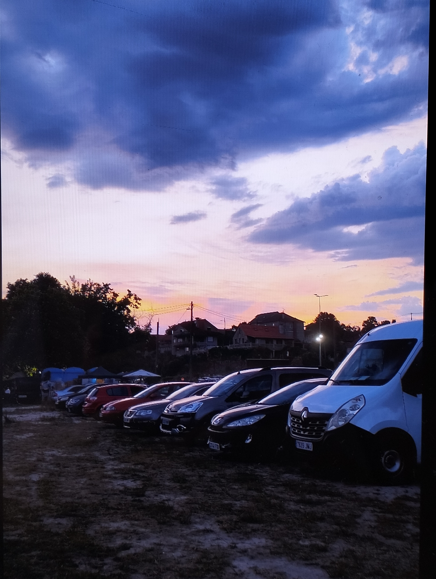 The image depicts a parking lot filled with cars and a white van, possibly at an outdoor event, as indicated by tents and people in the background. The scene is set at dusk, with a dramatic sky featuring dark, heavy clouds transitioning to hues of pink, orange, and purple near the horizon. Silhouetted trees and buildings are visible against the colorful sky. The ground appears to be dirt or gravel. A license plate on the white van is partially visible, showing the characters "7839 JW". The overall mood is tranquil, with the vibrant sunset creating a beautiful backdrop to the parked vehicles.
