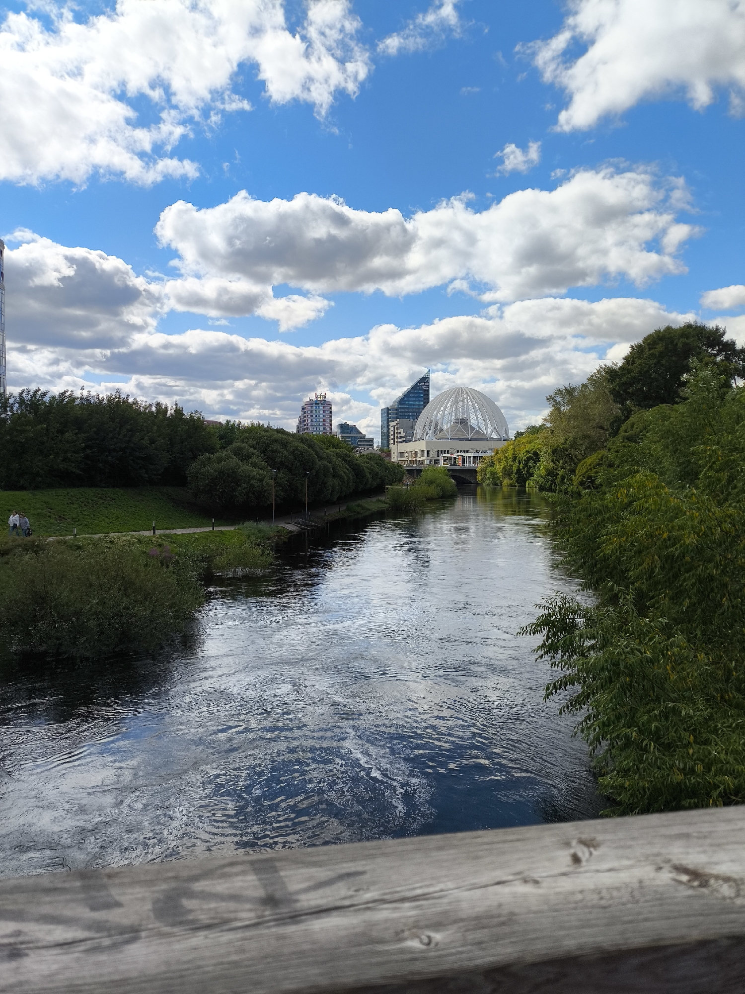 The image captures a wide river flowing through a city, with lush green trees lining the banks. In the background, modern buildings, including a distinctive domed structure, punctuate the skyline under a bright blue sky dotted with fluffy white clouds. On the left bank, a pathway with lampposts follows the river, and two individuals are visible walking along it. The foreground shows a wooden railing, suggesting the photo was taken from a bridge or elevated walkway. This scene is located on the Iset River in Yekaterinburg, Russia, with the domed building being the Yeltsin Center.