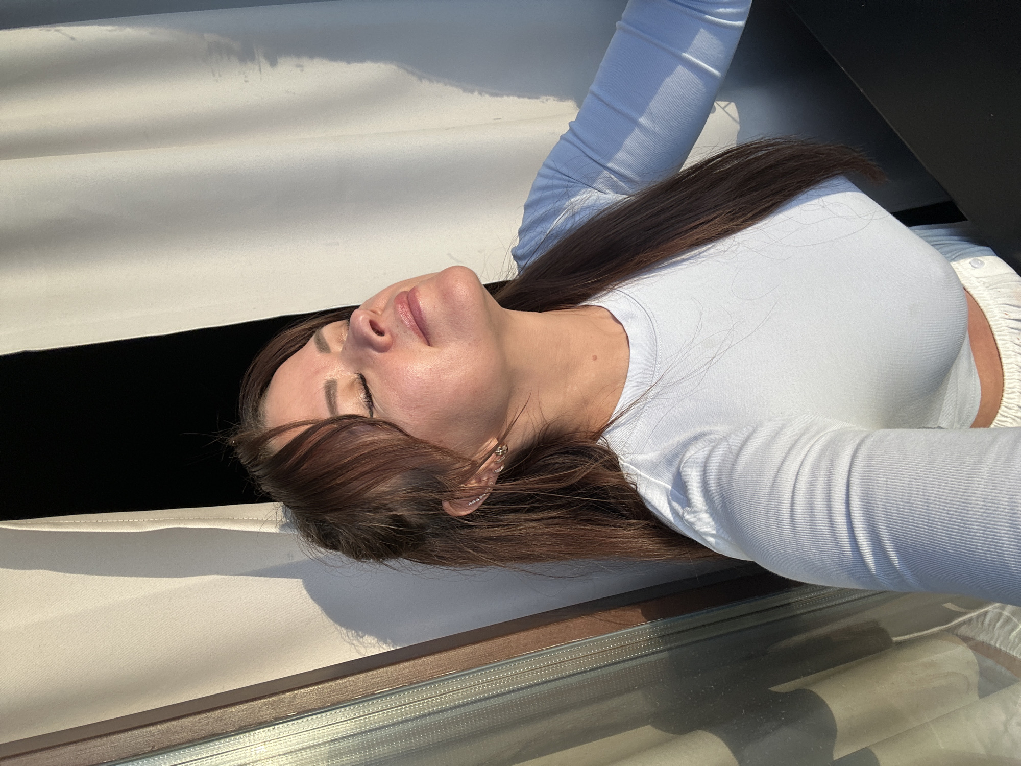 A young woman with long, brown hair is lying down, her head tilted back and her eyes closed. She is wearing a light blue long-sleeved top. The scene appears to be outdoors, with bright sunlight casting shadows. The background consists of a white textured surface and a dark, possibly black, area. There are also glimpses of what looks like a window or a reflective surface in the foreground. The woman has on subtle makeup, and a delicate earring is visible on her left ear. The overall mood seems relaxed and peaceful, perhaps suggesting a moment of repose or enjoying the sun. The image is taken from a low angle, emphasizing the woman's face and upper body.