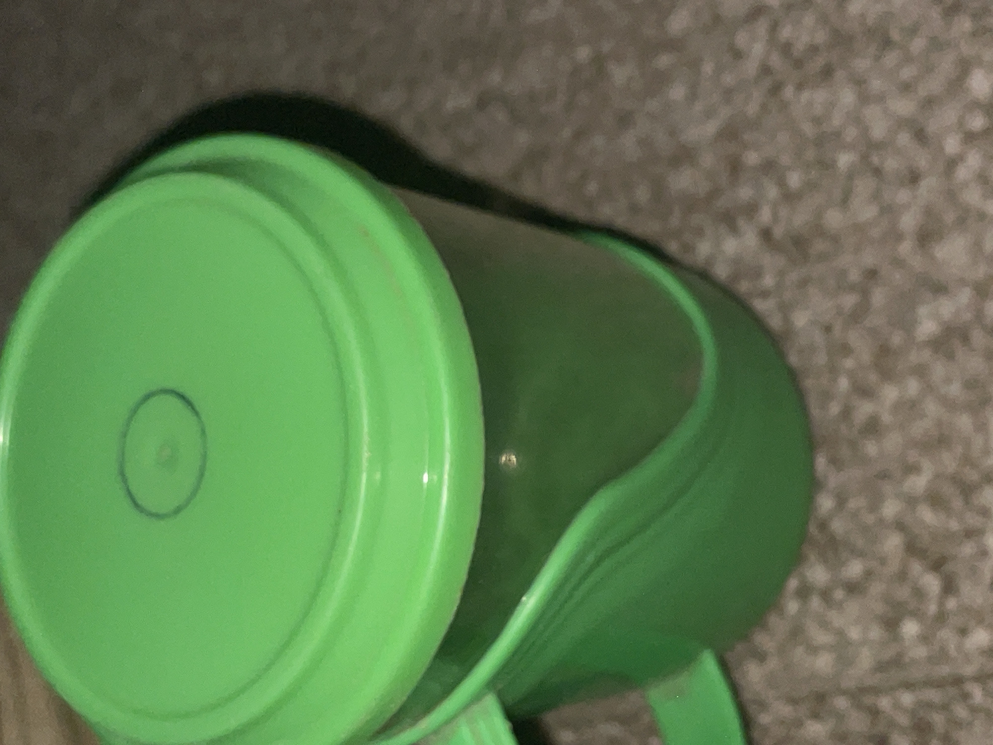 The media file shows a close-up, overhead view of a bright green plastic cup, possibly a child's training cup, lying on its side. The cup has a wide rim and a slightly translucent body, revealing a darker green interior. A faint, hand-drawn blue circle is visible on the exterior of the cup's flat top surface, with a small smudge or imperfection at its center. The background consists of a textured, neutral-colored surface, likely carpet or a similar fabric. The lighting appears to be artificial, casting shadows that suggest the cup is indoors. There is no visible text in the image. The setting is implied to be Jalingo, Nigeria, but this information is not visually derivable from the image itself. No people are visible, and the activity is static, simply depicting the object.