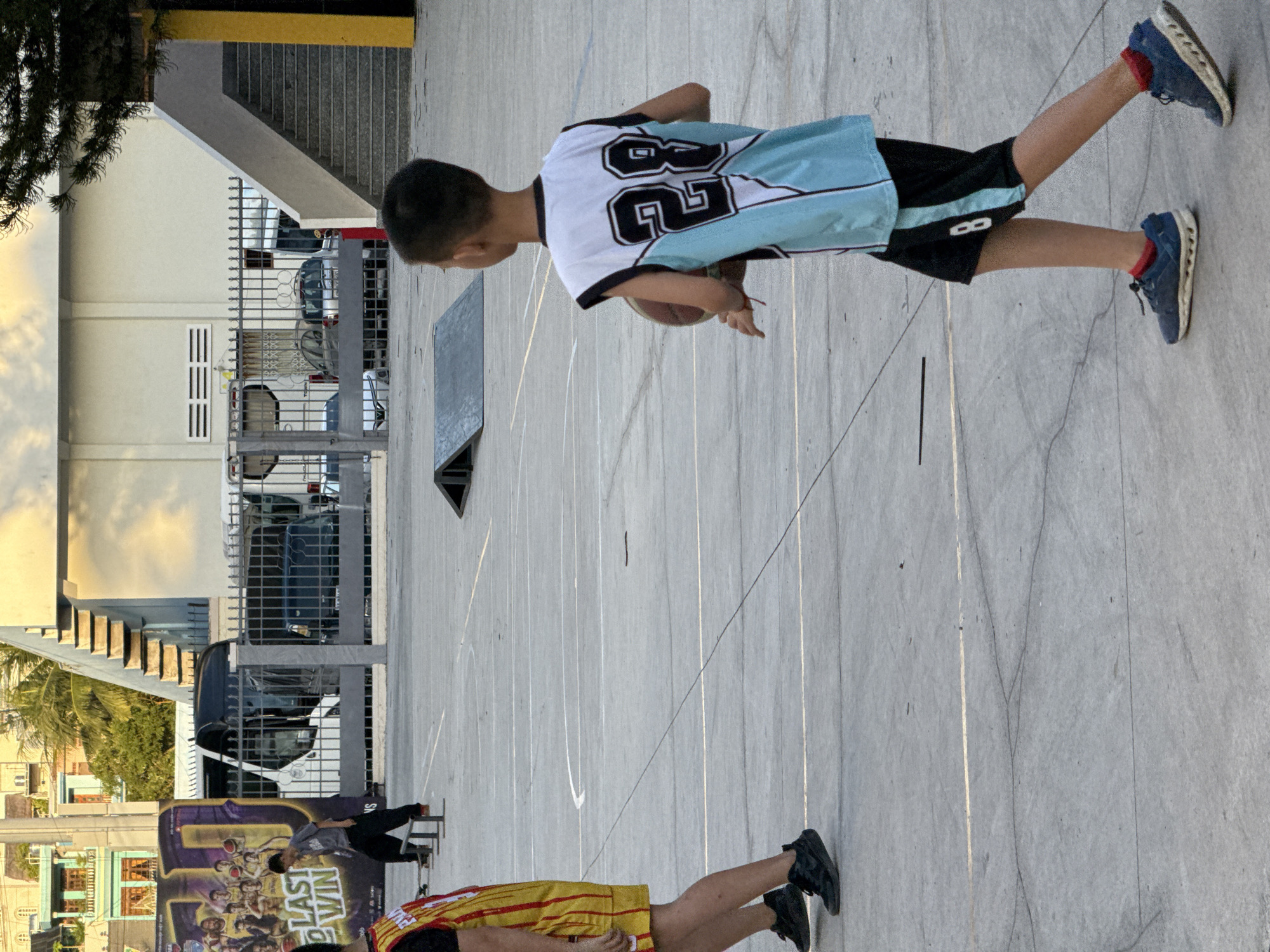 The image depicts an outdoor basketball court in Nha Trang, Vietnam, with an aged concrete surface marked by white lines and cracks. In the foreground, a young boy wearing a light blue and white basketball jersey with the number "28" on the back and black shorts with a white stripe is holding a basketball. He has red socks and blue athletic shoes. To the left, another person is partially visible, wearing a yellow and red striped basketball jersey and black sneakers.
In the background, a large banner with the text "LAST WIN" and images of basketball players is displayed. Adjacent to the banner is a bus, and beyond that, a fence separates the court from a street where cars are visible. Buildings with balconies and a staircase leading to an upper level are also present. Two skate ramps are positioned on the court. The scene appears to be during daylight hours, with the sky visible at the top of the frame.
