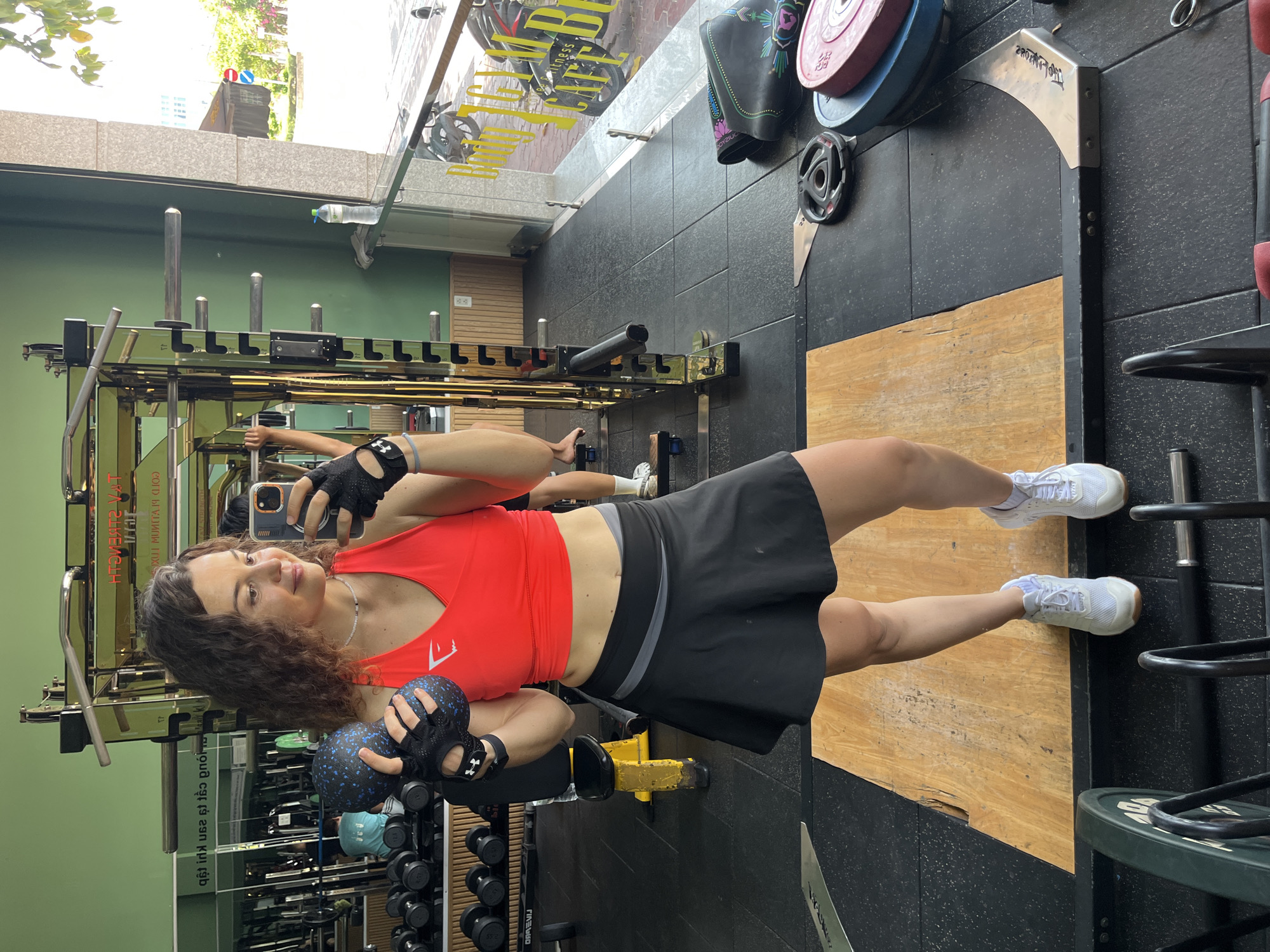 In Phan Thiết, Vietnam, a woman stands in a gym, taking a mirror selfie. She is wearing a bright red crop top, a black skirt, and white athletic shoes. She has dark, curly hair and is holding a blue and black dumbbell in her left hand and her mobile phone in her right. She is wearing black fingerless gloves. The gym environment is visible behind her, with weight racks, dumbbells, and gym equipment. Through a large window to her right, a street scene with trees and buildings is visible. The gym floor is black rubber, with a wooden platform in the foreground.