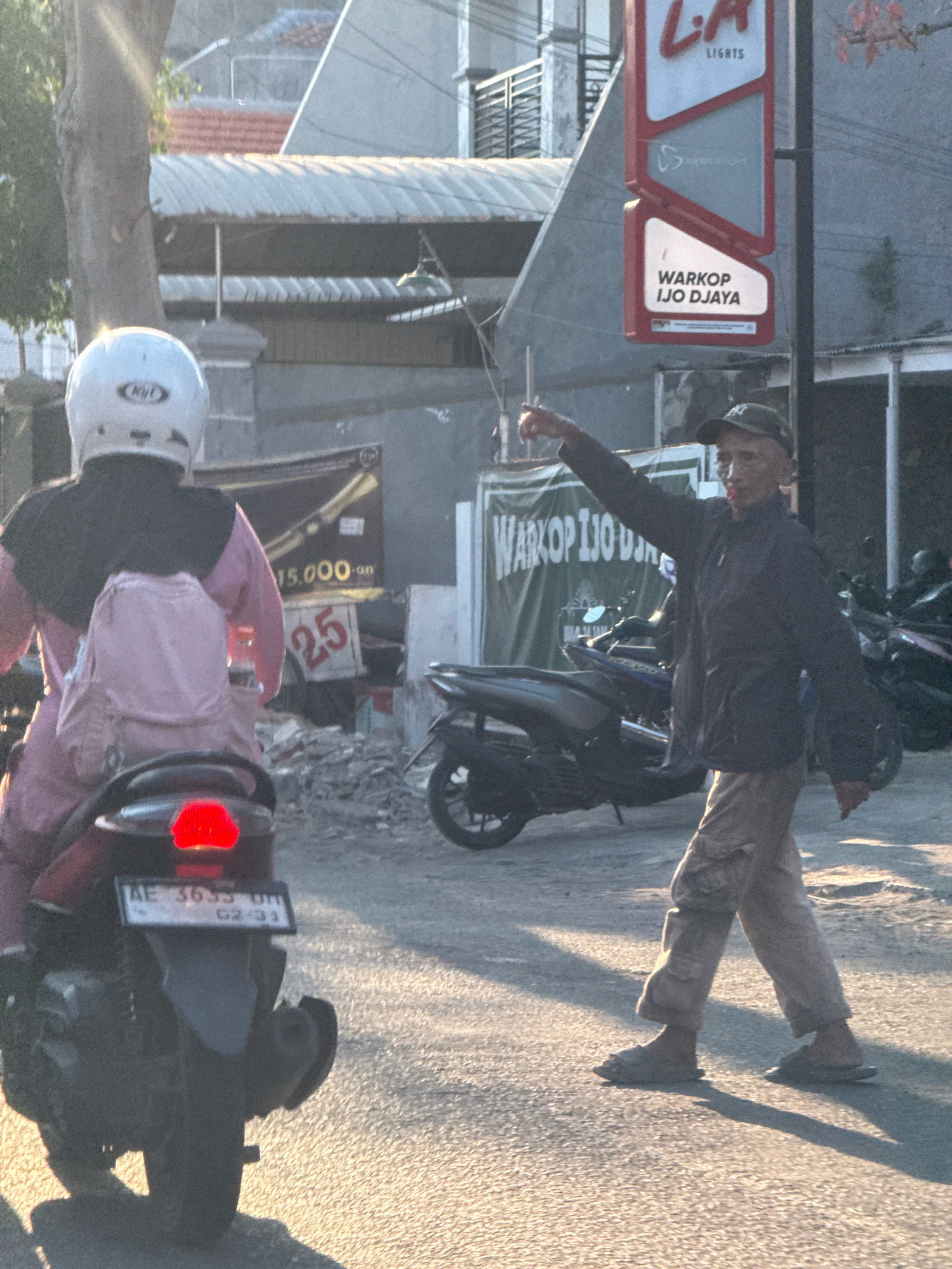 In Surabaya, Indonesia, on a street bathed in sunlight, a person wearing a white helmet and pink clothing is riding a motorcycle, facing away from the camera. Behind them, a man in a dark jacket and light-colored pants is walking and gesturing with his right hand. A sign with "L.A. LIGHTS" and "WARKOP IJO DJAYA" is visible in the background. Several motorcycles are parked on the side of the road. A building with a corrugated metal roof is also present. The scene appears to be a street in an urban area.