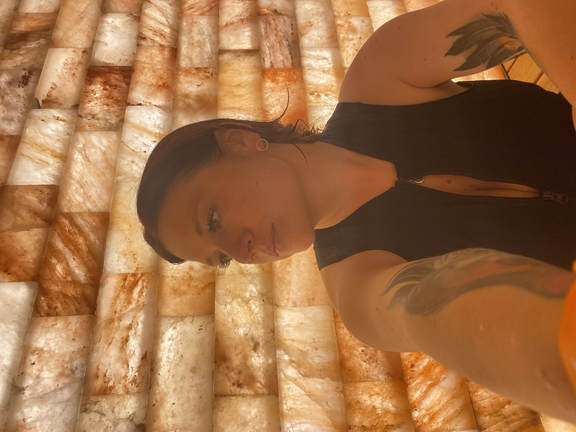 The image shows a woman in a salt cave. The walls are made of salt blocks, which are lit from behind, giving them an orange and white glow. The woman is wearing a black sleeveless top with a zipper. She has tattoos on her arms. She is looking to her left, with her head slightly tilted. The salt cave appears to be an indoor space, likely a spa or wellness center, located in Votkinsk, Russia. There are no other people or objects clearly visible in the image. The lighting is warm and diffused.