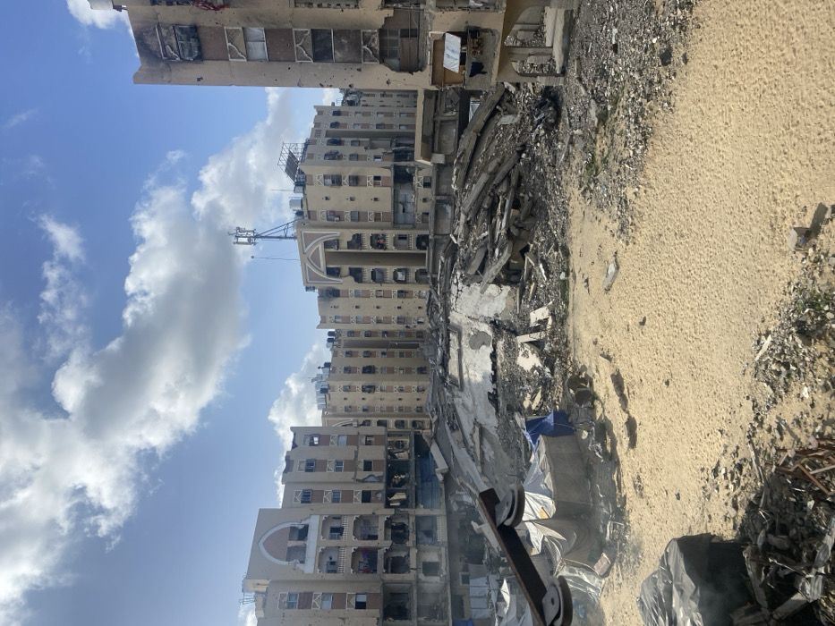 A damaged building complex stands under a partly cloudy sky. Evidence of severe destruction is present, including collapsed walls, burnt areas, and debris scattered across the ground. A dirt or sandy area is also visible near the building. A cell phone tower is on top of the buildings. The scene suggests a conflict or disaster area.