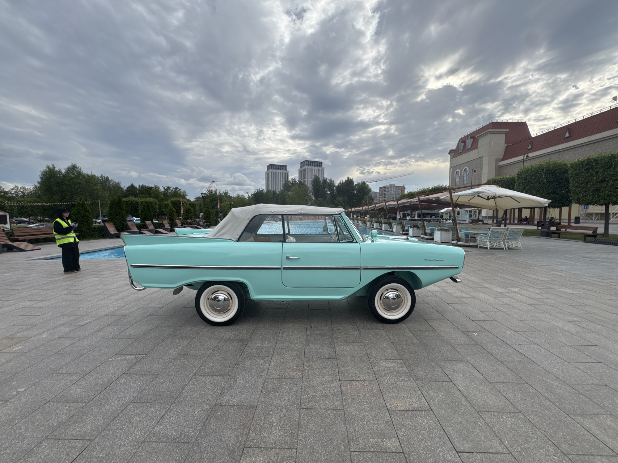 A turquoise amphibious vehicle, likely a Schwimmwagen or similar, is parked on a grey stone-tiled pedestrian area next to a bright blue swimming pool. The vehicle has a light-colored convertible top up, white-walled tires with chrome hubcaps, and a distinctive finned rear. To the left, a person wearing a yellow safety vest and black pants stands near the pool. In the background, there are trees, lounge chairs, tables with umbrellas, and several modern buildings, including two tall, light-colored towers. The sky is overcast with dramatic clouds. The scene appears to be an outdoor recreation or event area in Moscow, Russia, given the architectural style of the buildings and the presence of a pool.
