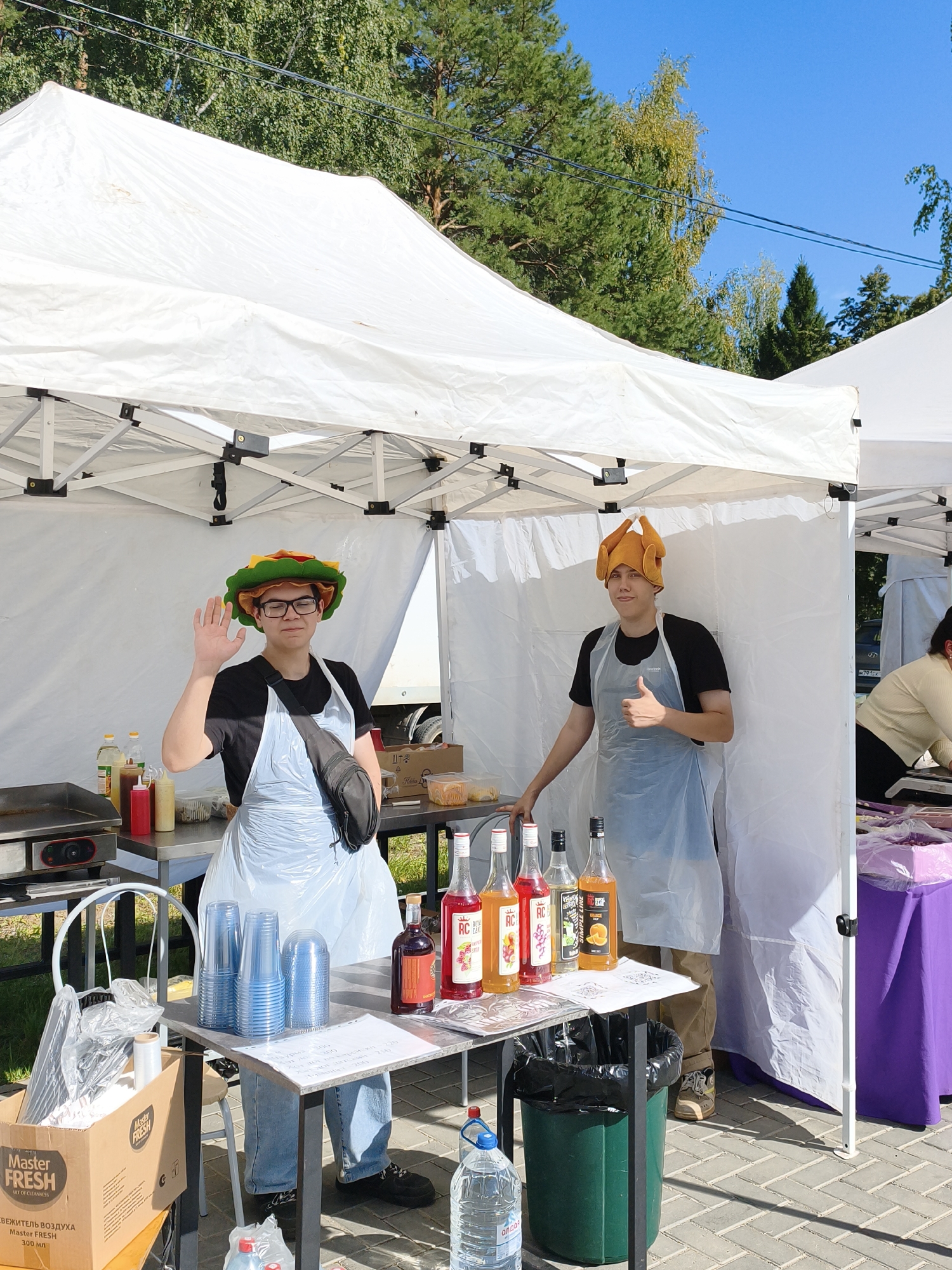 A medium shot captures two young men, presumably employees, standing under a white canopy tent at an outdoor market stall. The man on the left, wearing glasses, a black t-shirt, and a light blue plastic apron over jeans, waves at the camera with his left hand. He sports a novelty hat shaped like a burger. To his right, the second man, in a black t-shirt, light blue plastic apron, and light brown pants, gives a thumbs-up with his right hand. He wears a novelty hat resembling a roasted chicken.
The stall is set up on a paved area, with a checkered pattern visible on the ground. A metal table in the foreground displays various items. To the left, a flat-top grill and condiment bottles are visible. The table holds several stacks of blue plastic cups, bottles of colored syrup, and a handwritten menu. A black trash can sits beside the table, and a large water bottle is on the ground. A cardboard box with "Master Fresh" branding is placed near the table. In the background, trees and a bright blue sky are visible. To the right, another stall with a purple tablecloth and a woman in the background suggests a bustling market environment. The scene is indicative of a food or drink vendor operating at an outdoor event.