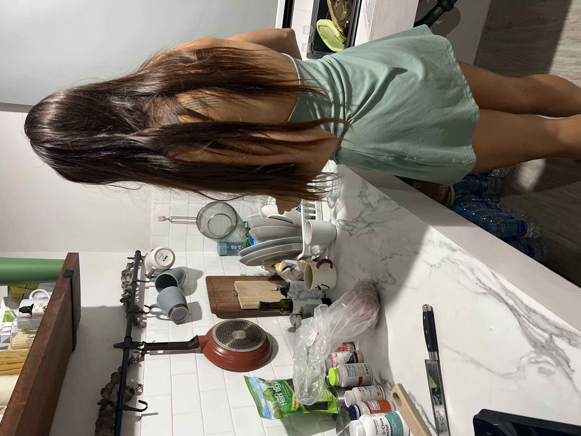 An indoor, eye-level shot captures a person from behind, with long, dark hair flowing over their shoulders and back. They are wearing a light green, sleeveless dress and appear to be standing at a kitchen counter, possibly engaged in food preparation or cleaning. A tattoo is visible on their left shoulder/upper arm.
The kitchen counter, featuring a white marble-like pattern, is laden with various items. To the person's left, there's a black metal rack with several ceramic mugs hanging, and a red frying pan resting on the countertop. A bag labeled "Heritage Nature's" is visible, along with multiple supplement bottles, some clearly showing "Fairhaven D-Chiro Inositol" and "D-Chilo". A large kitchen knife lies on the counter near these bottles. Directly in front of the person, a sink area is implied, with dirty dishes stacked in a drying rack. A wooden cutting board is also on the counter. Below the counter, stacks of clear plastic water bottles are visible. The walls feature white square tiles. The lighting suggests an indoor setting, likely evening or artificial light.
Visible text includes: "D-CHILO", "Fairhaven D-Chiro inositol", "N", "Heritage Nature's", "FOR SHACKING & BAN", and "SS". The scene is located in บ้านบางรักษ์, Thailand.