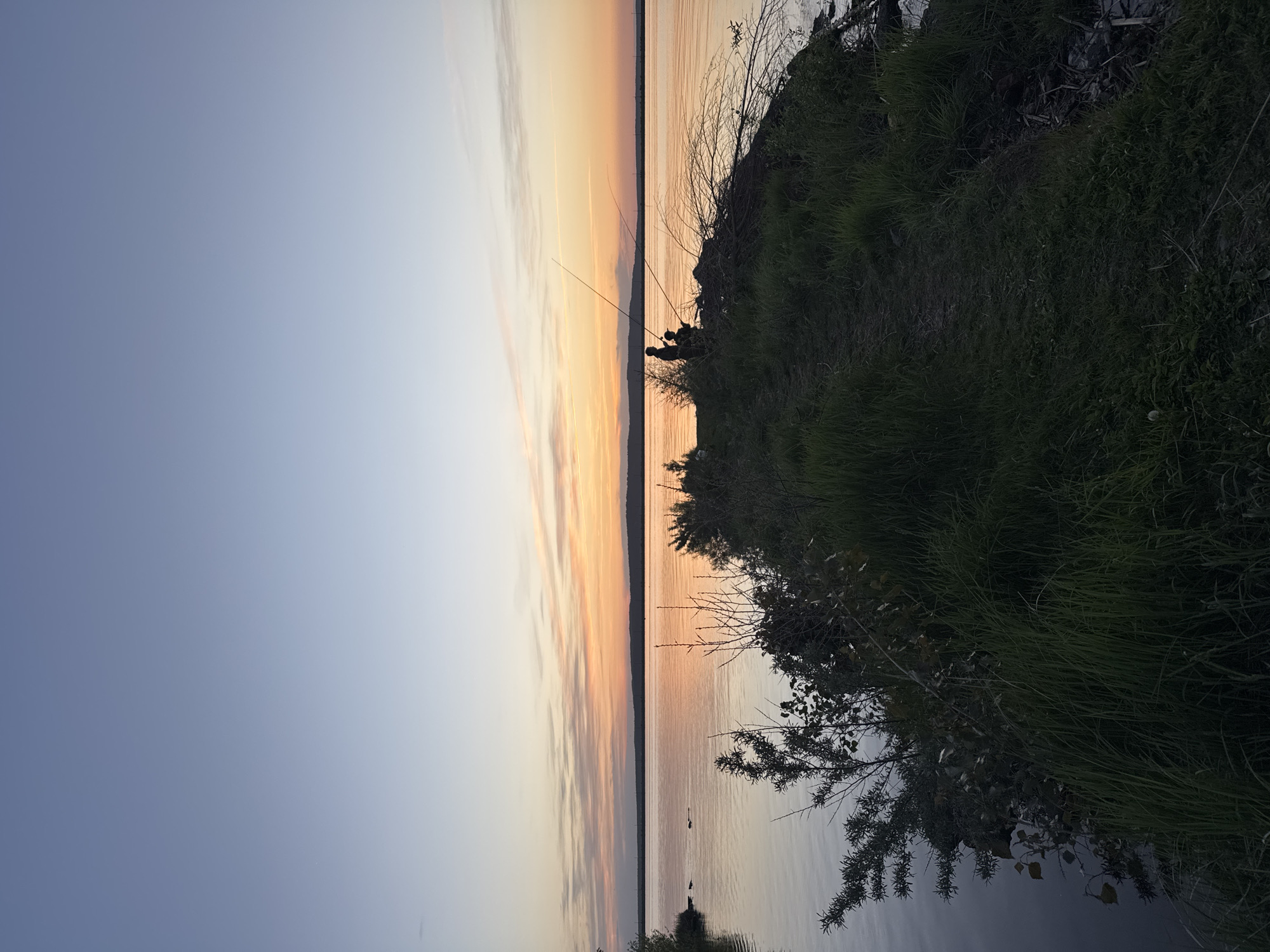 The image depicts a sunset over the Kama River in Chaikovsky, Russia. Silhouetted figures, possibly fishermen, are positioned on the riverbank with fishing rods extending towards the water. The sky displays a gradient of colors from light blue to orange, reflected in the calm water. Vegetation, including grass and small trees, lines the riverbank.
