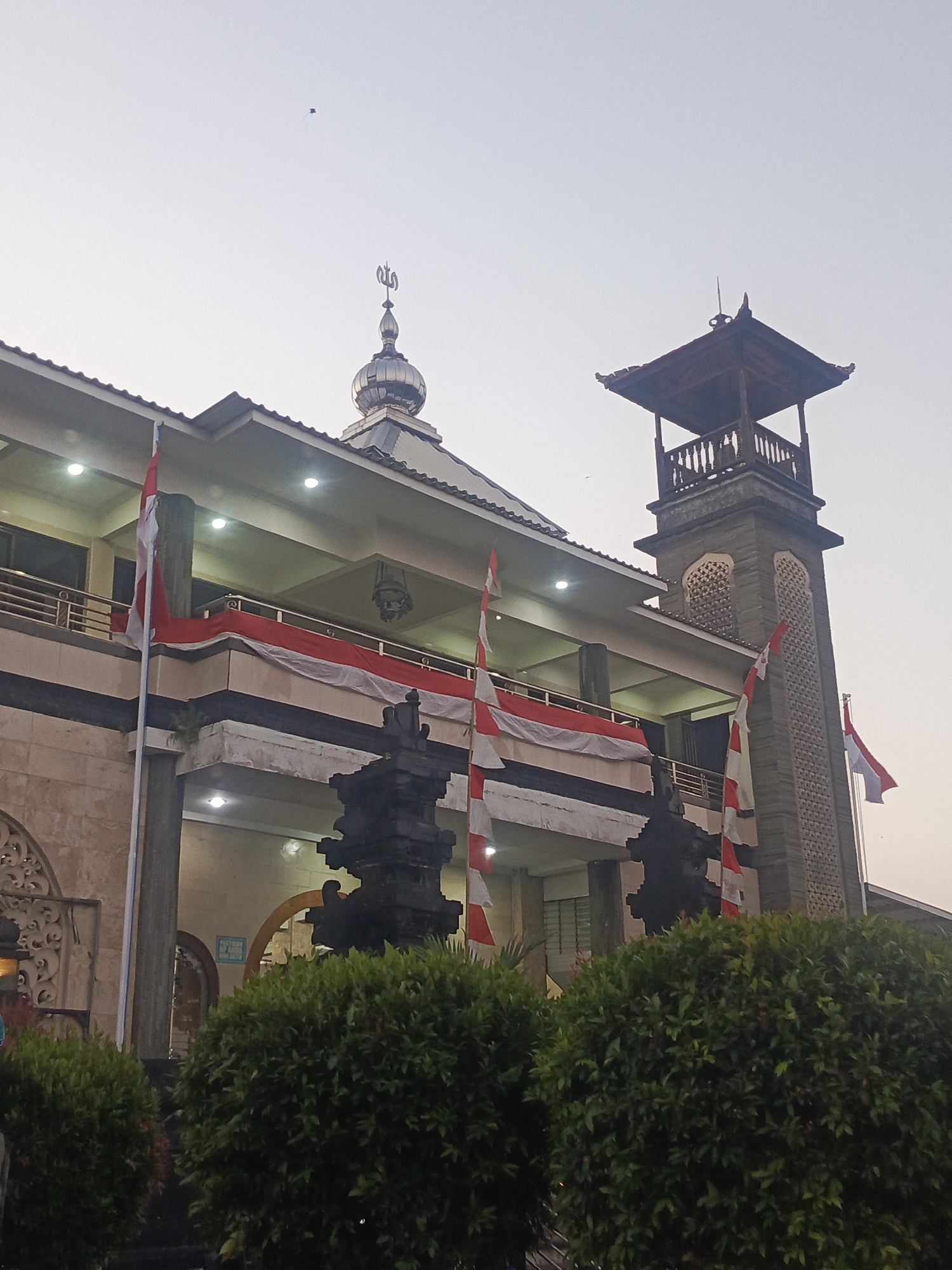 The image shows a mosque with Indonesian flags displayed, located in the city of North Kuta, Indonesia. The mosque has a dome with an Islamic inscription and a tall, square tower with an ornate wooden structure at the top. Red and white striped flags are hung along the facade of the mosque and from flagpoles. Lush green bushes are visible in the foreground, partially obscuring the lower level of the building. The sky is overcast. In the distance, a kite is visible flying in the sky.