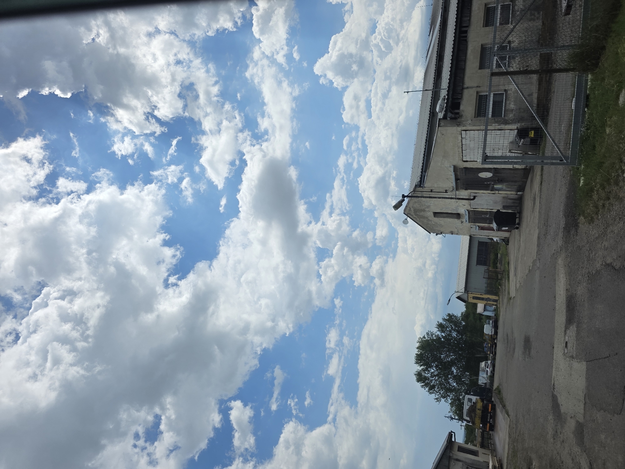 The image shows a view of the sky with scattered clouds and part of an industrial or rural area. The sky is predominantly blue, filled with cumulus clouds of varying sizes and densities. In the foreground, a building with a utilitarian design and visible signs of aging is present, featuring a corrugated roof, several windows, and a metal gate. A paved road or street runs alongside the building, and there is vegetation near the edge of the road. A tree is visible further down the street, and there are parked vehicles in the distance. The location appears to be in a non-urban or outskirts area, suggestive of an industrial park or a rural setting. The image does not contain people, actions, or interactions. The location is Unknown, Unknown.