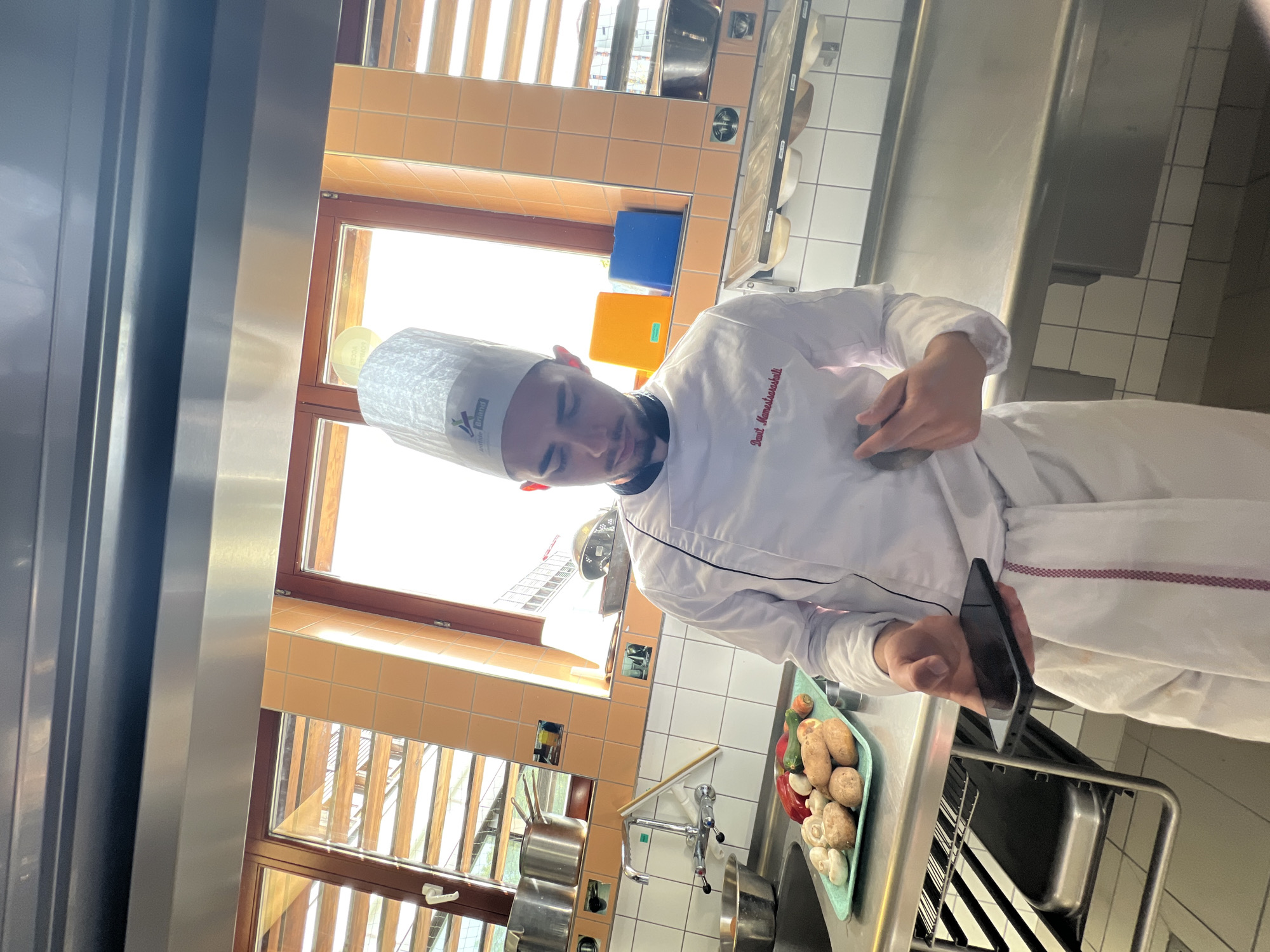 A person wearing a chef's uniform and hat stands in a kitchen, holding a black smartphone in their right hand. They are looking down at the phone. A tray containing mushrooms, potatoes, zucchini, and red peppers is on the counter beside them. In the background, there is a window with blinds, stainless steel countertops and a sink. The location is possibly a restaurant or catering kitchen in Schiltigheim, France, based on the writing on the chef's hat.