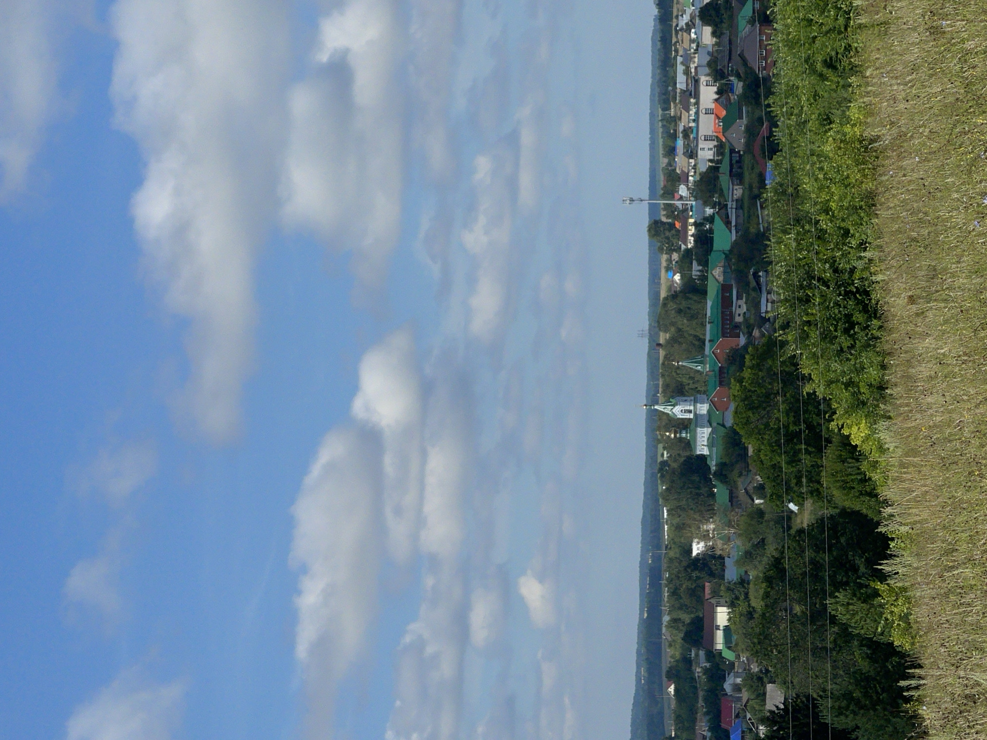 The image depicts a wide view of a rural landscape on a sunny day with a blue sky filled with scattered white clouds. In the foreground, a dry, grassy hill slopes upward. In the mid-ground, a small town is visible, characterized by numerous houses with green and red roofs, interspersed with trees and a prominent white church with a green dome and spire. A cell tower stands tall on the horizon. The background consists of a distant treeline and rolling hills under the vast sky. The scene appears to be a tranquil day in the Russian countryside, possibly the city of Perevoznoe, Russia. There are no people visible in the image.