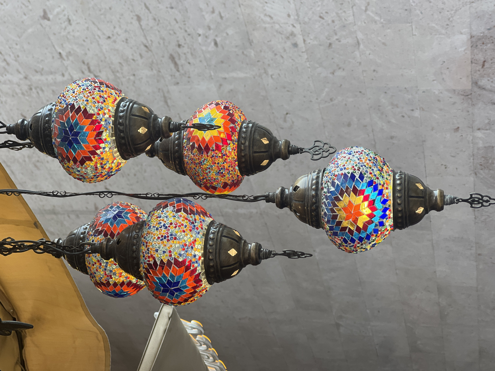 The image shows a low-angle shot of four hanging Turkish lamps. The lamps have a metal frame and are decorated with colorful mosaic tiles. The lamps are suspended from a metal chain against a background of a grey ceiling and awning. The location is possibly Yerevan, Armenia, given the prevalence of Turkish cultural elements.