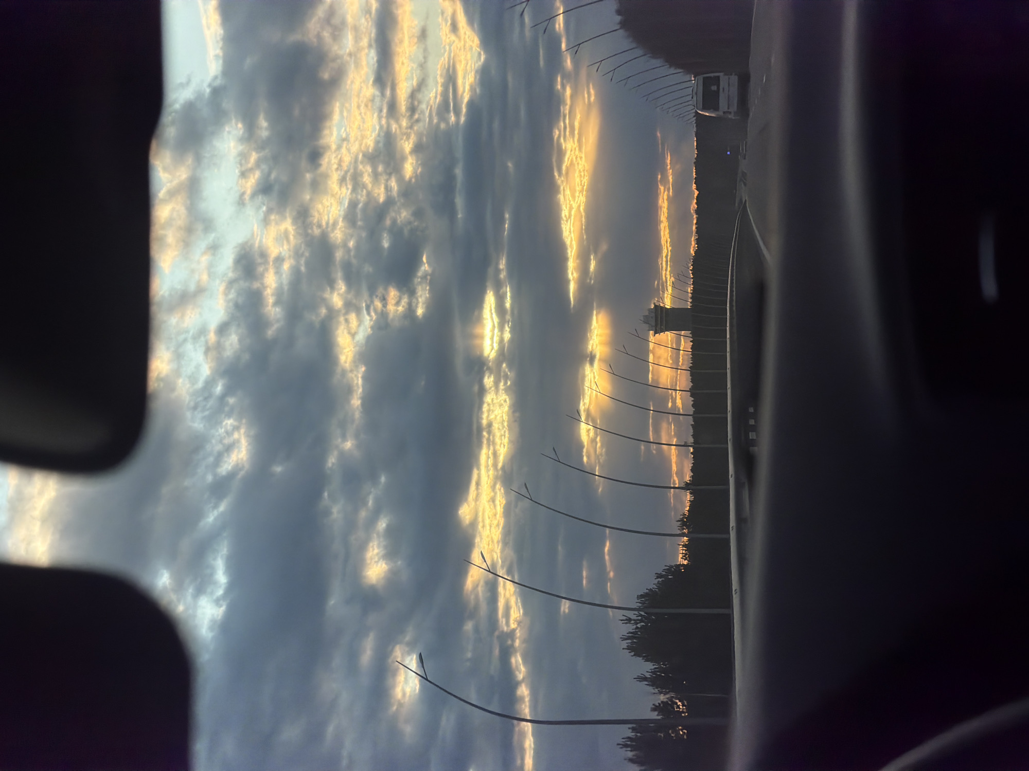 The image depicts a view from inside a vehicle, likely a car, during what appears to be a sunset. The sky is filled with clouds, some darker gray and some illuminated with a golden hue. There are a number of streetlights visible, curving over a highway or large road. In the distance, there appears to be an overpass. The trees border the edge of the road. The scene is taken during a sunset or sunrise. Based on the prompt, it is located in Krasnoyarsk, Russia.