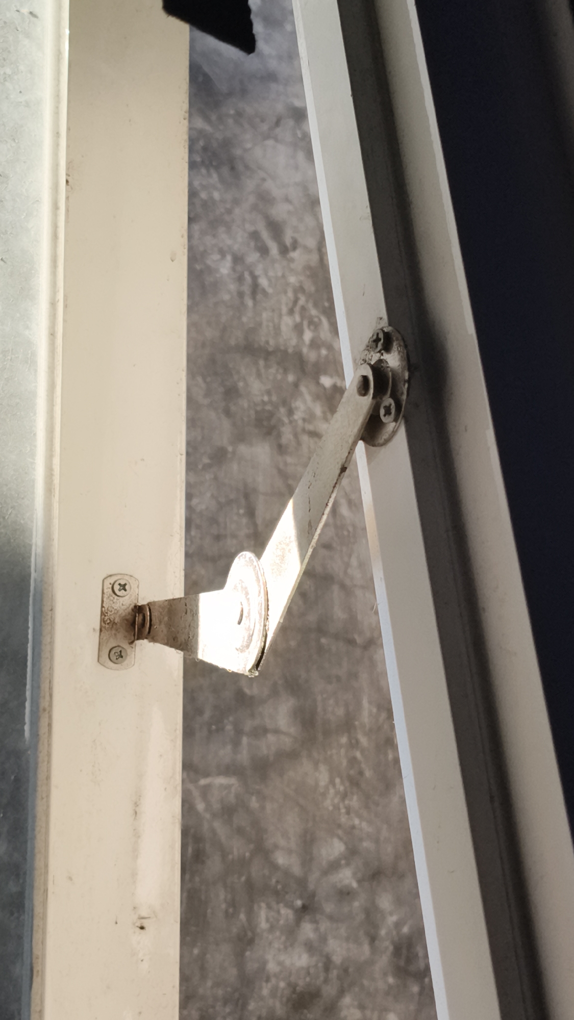 The image shows a close-up of a window hinge mechanism. A cream-colored window frame is visible on the left, with a metal hinge attached by screws. The hinge extends outwards and is connected to another part of the window frame, which is also cream-colored and appears to be a window sash. The hinge itself is made of metal and shows signs of wear and tear, with some rust and dirt. A portion of the window opening is visible behind the hinge, revealing a textured, grey surface, possibly a wall with a concrete finish. Sunlight casts a strong shadow on the hinge and the surrounding frame, highlighting the details of the mechanism. The scene is likely indoors, near a window, and the overall impression is of an aging but functional window hardware. The location in Kerep Kidul, Indonesia, suggests a tropical or sub-tropical climate, which might contribute to the wear on the metal components due to humidity and exposure to elements.