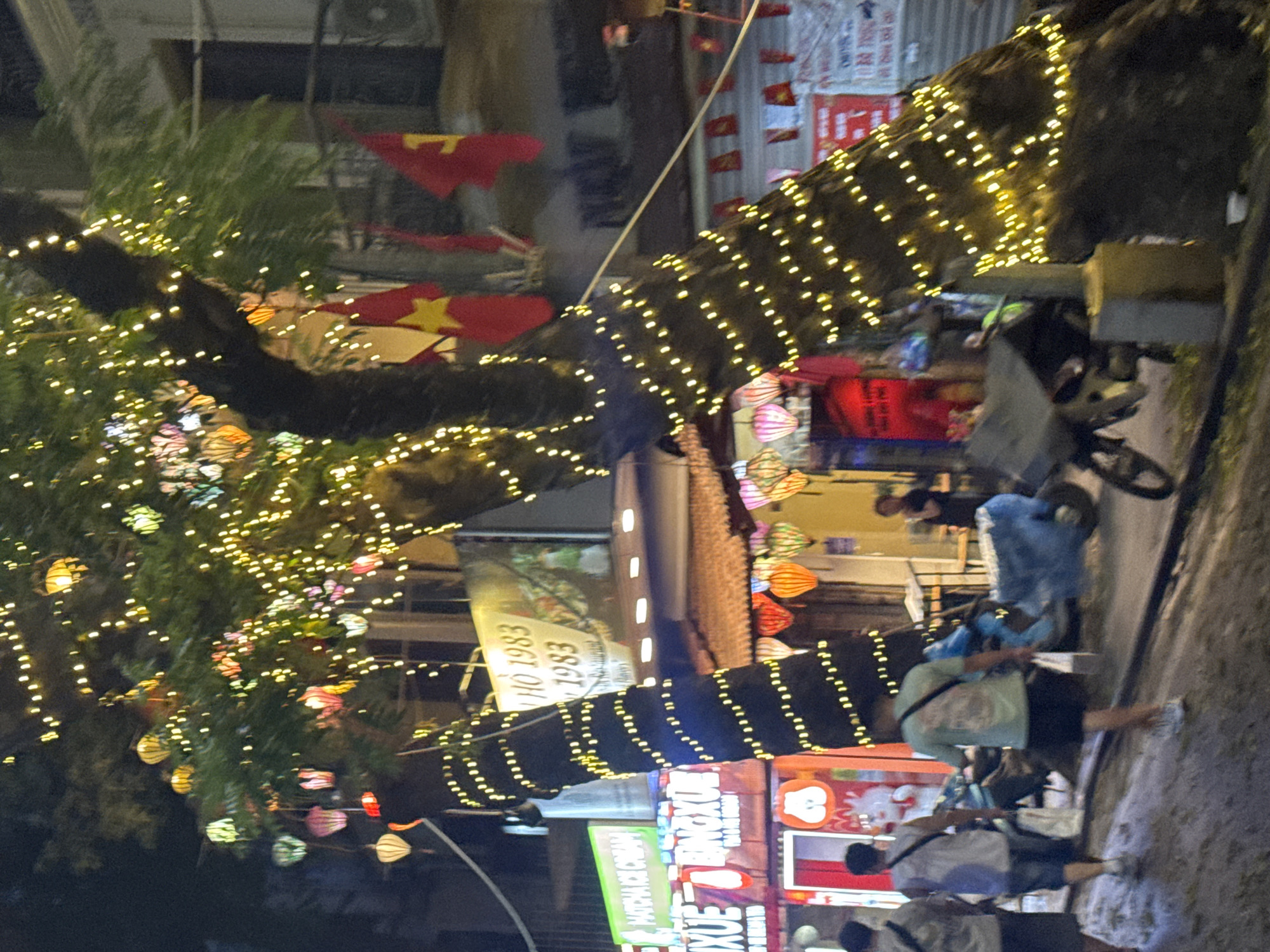 The image depicts a street scene in Hà Nội, Vietnam, at night. The view is angled. Strings of lights are wrapped around tree trunks. Lanterns hang from the trees. Vietnamese flags are visible in the background. People walk along the sidewalk. Shops and restaurants line the street. A sign reads "Matcha Ice Cream".
