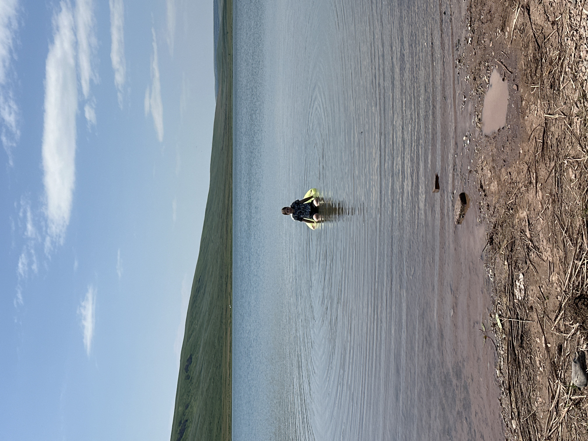 A young person sits on a bright yellow flotation device in a large, calm body of water. The water has ripples spreading outwards from the flotation device, indicating movement. The person is wearing a dark t-shirt with a white pattern and dark shorts. They are facing the camera. The water's edge in the foreground is rocky and muddy, with some dry vegetation scattered on the ground. In the background, a gently sloping, green grassy hill rises to meet a blue sky with scattered white clouds. The scene appears to be a natural outdoor setting, likely a lake or reservoir, under a sunny sky. There is no visible indication of the city of Novonikolaevka, Russia in the image.