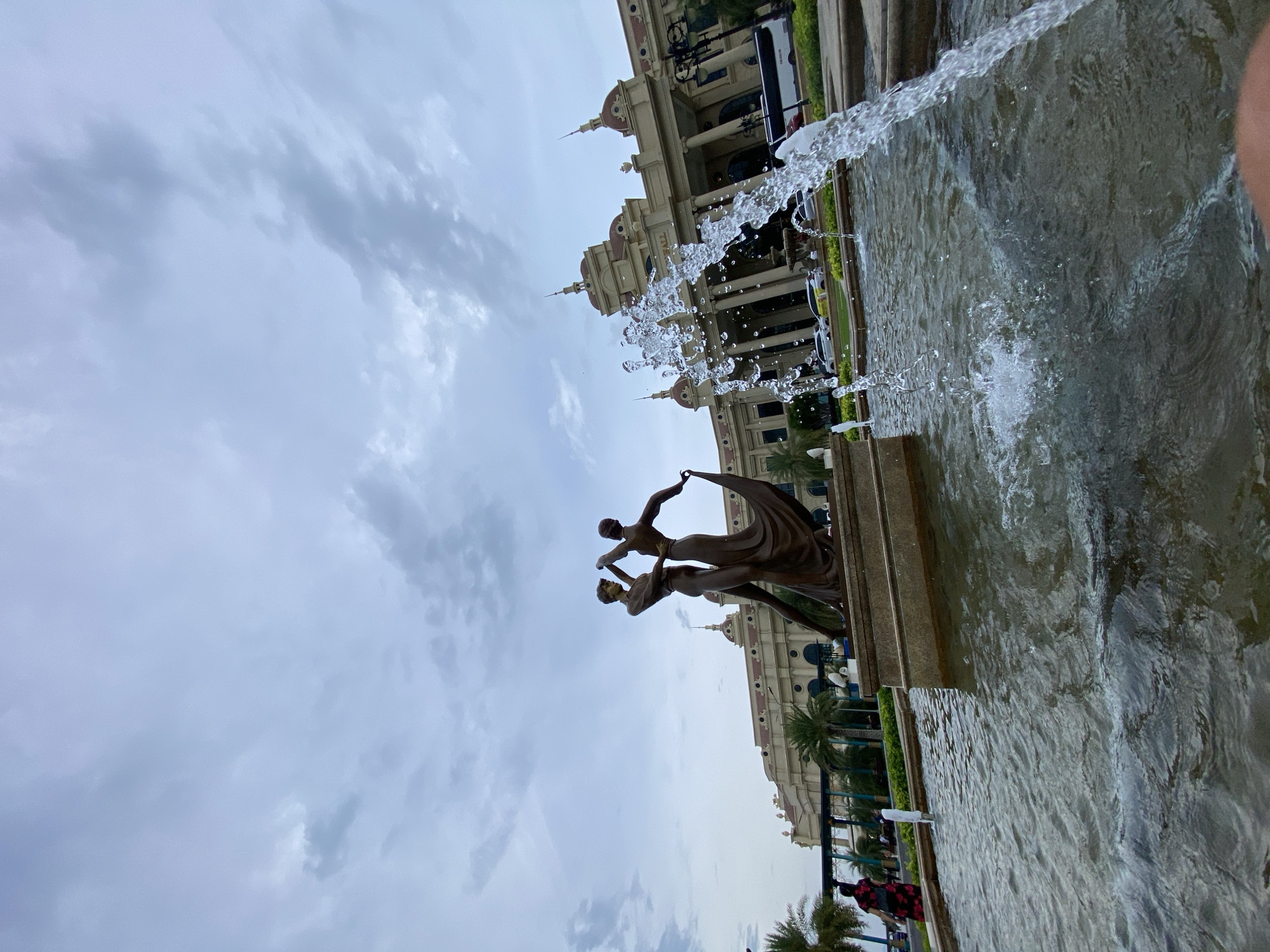 The image captures an outdoor scene in Nha Trang, Vietnam, featuring a bronze statue of two dancers in the foreground, positioned in front of a large, ornate building with classical architectural elements, including columns and domes. A fountain with splashing water is prominently visible in the lower right portion of the frame, adding dynamic movement to the scene. In the background, palm trees and other greenery are present, suggesting a tropical or subtropical climate. The sky is overcast with scattered clouds. Several cars and a bus are visible parked in front of the building, and a few individuals can be seen walking in the distance. The overall impression is of a public space with both artistic and architectural significance.
