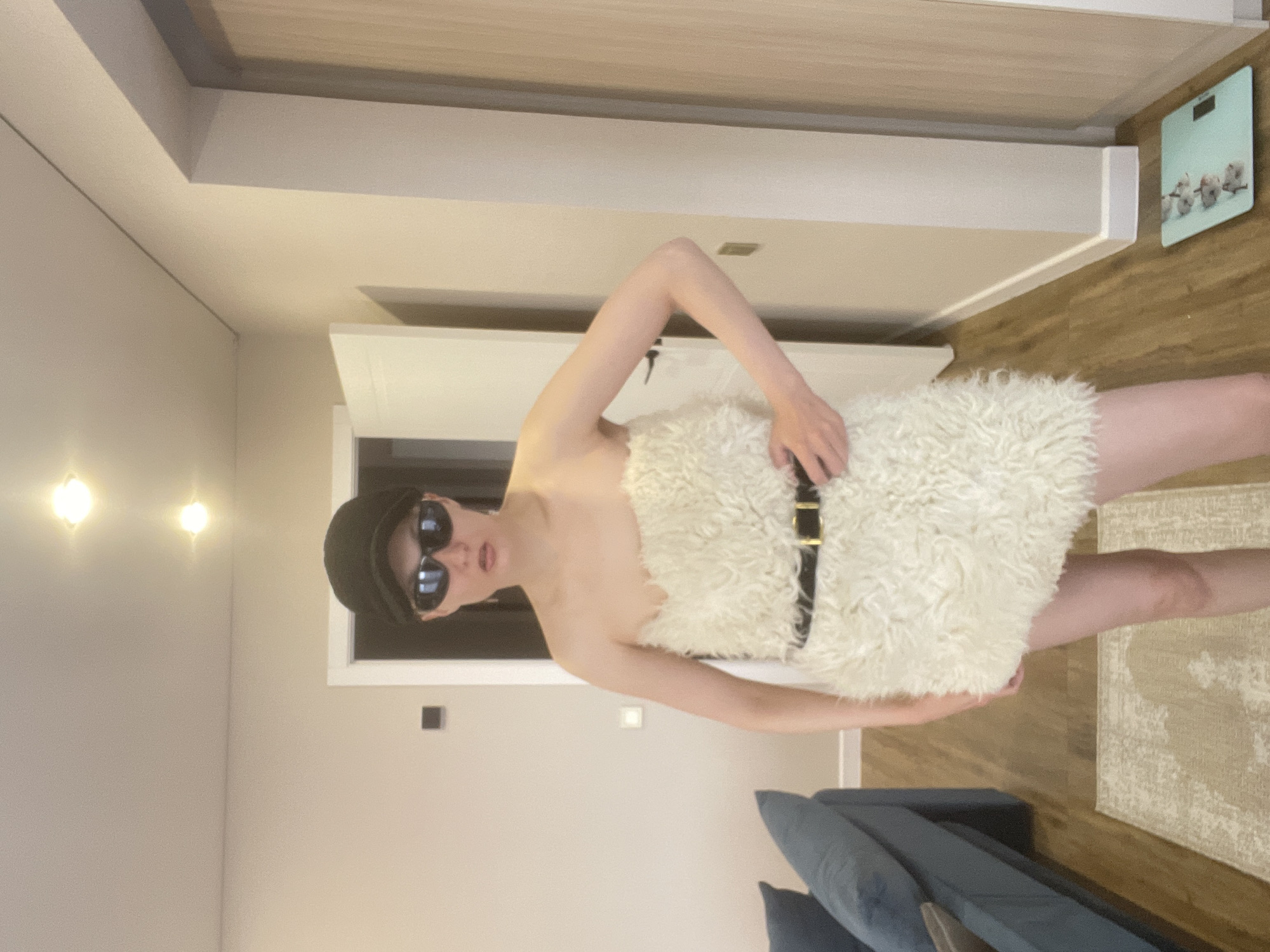 The image appears to be taken in an indoor setting, possibly a residence in Подшивалово, Russia, though location cannot be confirmed. A person, dressed in what appears to be a white, shaggy garment secured with a black belt, stands in the center of the frame. The individual is wearing a dark-colored hat and sunglasses. The background features a doorway, wall lighting, a bathroom scale, a rug, and a blue couch.