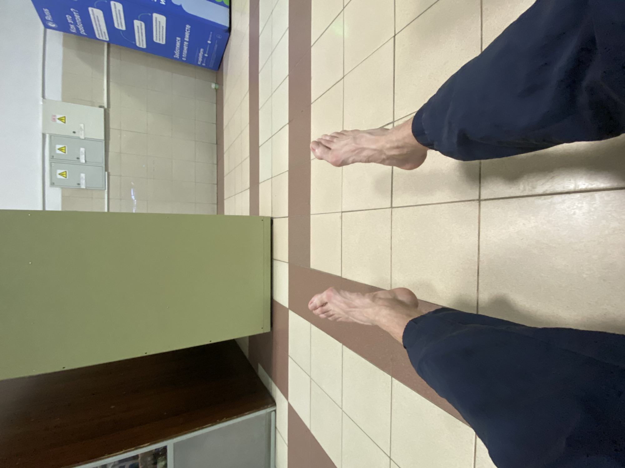 This image, rotated 90 degrees counter-clockwise, depicts a first-person view of a person resting indoors, with their bare feet and lower legs in dark trousers visible. The perspective suggests the person is sitting or lying down on a light-colored tiled floor, which features a pattern of light beige or off-white square tiles interspersed with wider, light brown striped sections.
In the foreground, to the left of the frame (which would be the bottom if upright), a light olive-green structure, possibly a bench or a low counter, partially obscures the view.
In the background, on what appears to be a white-tiled wall, three light grey electrical panels are mounted. Each panel features a yellow triangle warning symbol, indicating caution. To their right, a tall blue information stand or display is visible.
Visible text on the blue stand, in Russian, includes:
* "Russ" (likely a brand name or logo)
* "Как это работает?" (How does this work?)
* "Заботимся о планете вместе" (We care about the planet together)
* "ecoplatform" (a small text, possibly a website or platform name)
The indoor setting, tiled floor, electrical panels, and public information stand suggest a public or commercial building, such as a waiting area, shopping center, or transportation hub in Ryazan, Russia, as indicated by the Russian text. The lighting appears to be artificial, consistent with an indoor environment. No specific time of day or emotions are discernible.