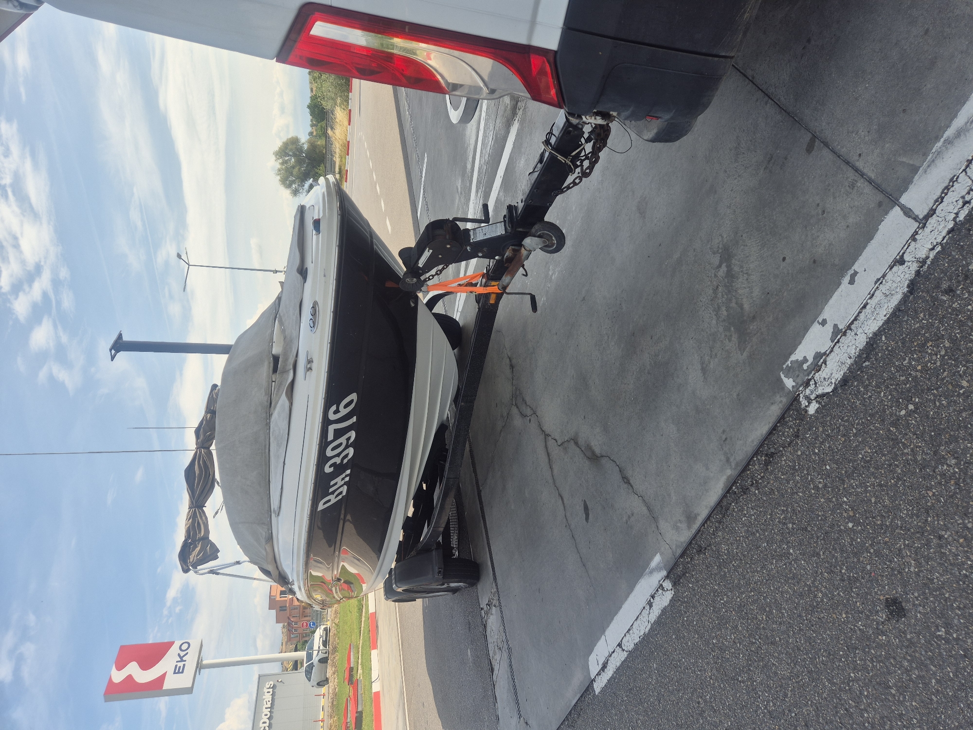 A white and black motorboat is parked on a trailer on a paved surface. The boat is covered with a grey tarp, and the registration number "BH 3976" is visible on its side. The trailer is attached to a white van, which is partially visible on the right side of the image. In the background, there is an EKO gas station sign and a McDonald's building. A white car is parked near the McDonald's. The sky is blue with some clouds. The scene is located in Chuchuligovo, Bulgaria.