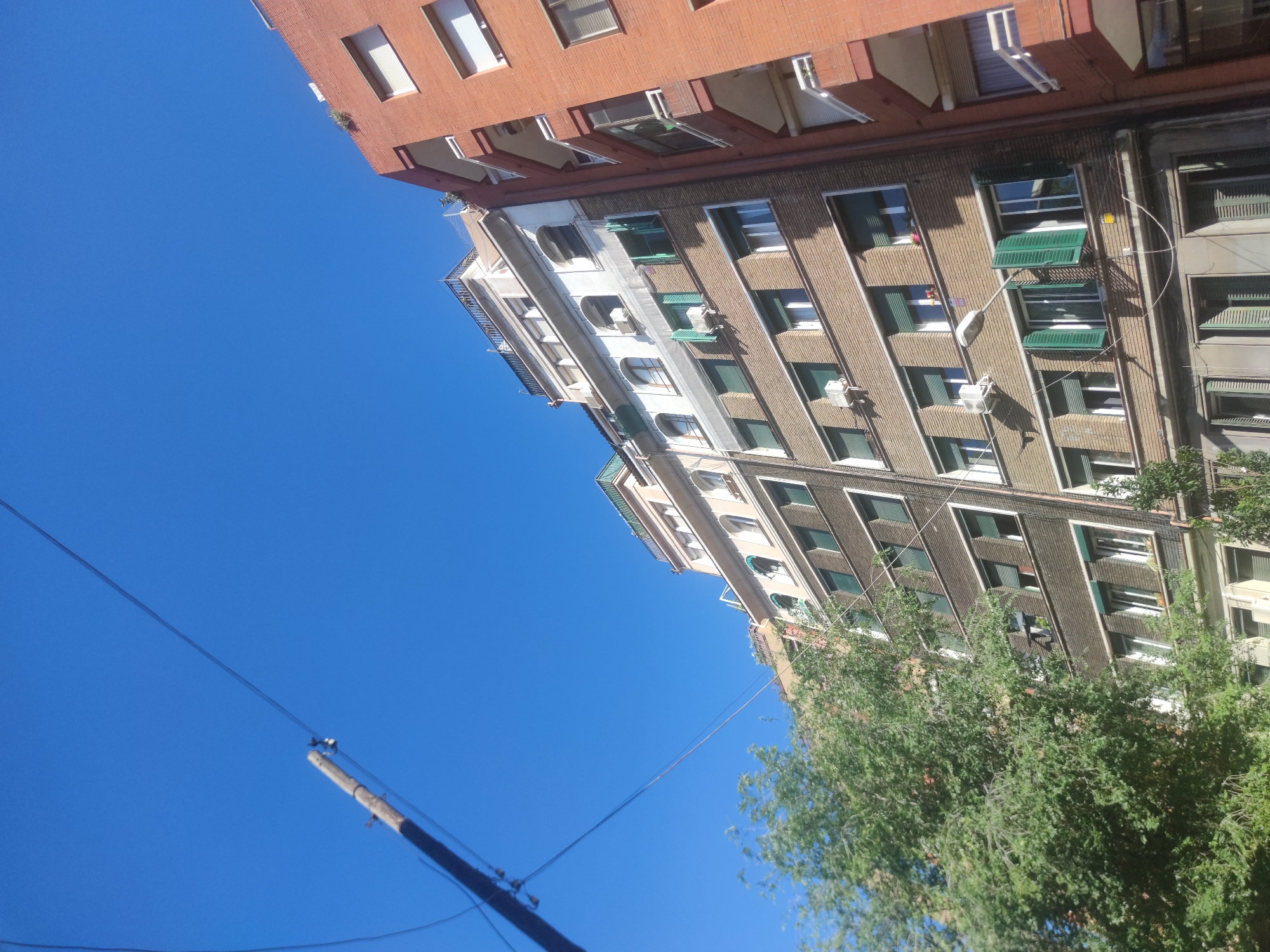 A low-angle shot of a street scene shows the sides of two multi-story buildings under a clear blue sky. The building on the left is made of red brick and has rectangular windows with white frames and awnings. The building next to it is composed of a white section and a dark-gray section with rectangular windows, some of which have green shutters. A utility pole with power lines runs from the lower left of the image towards the buildings. Trees with green foliage are visible on the right side of the buildings.