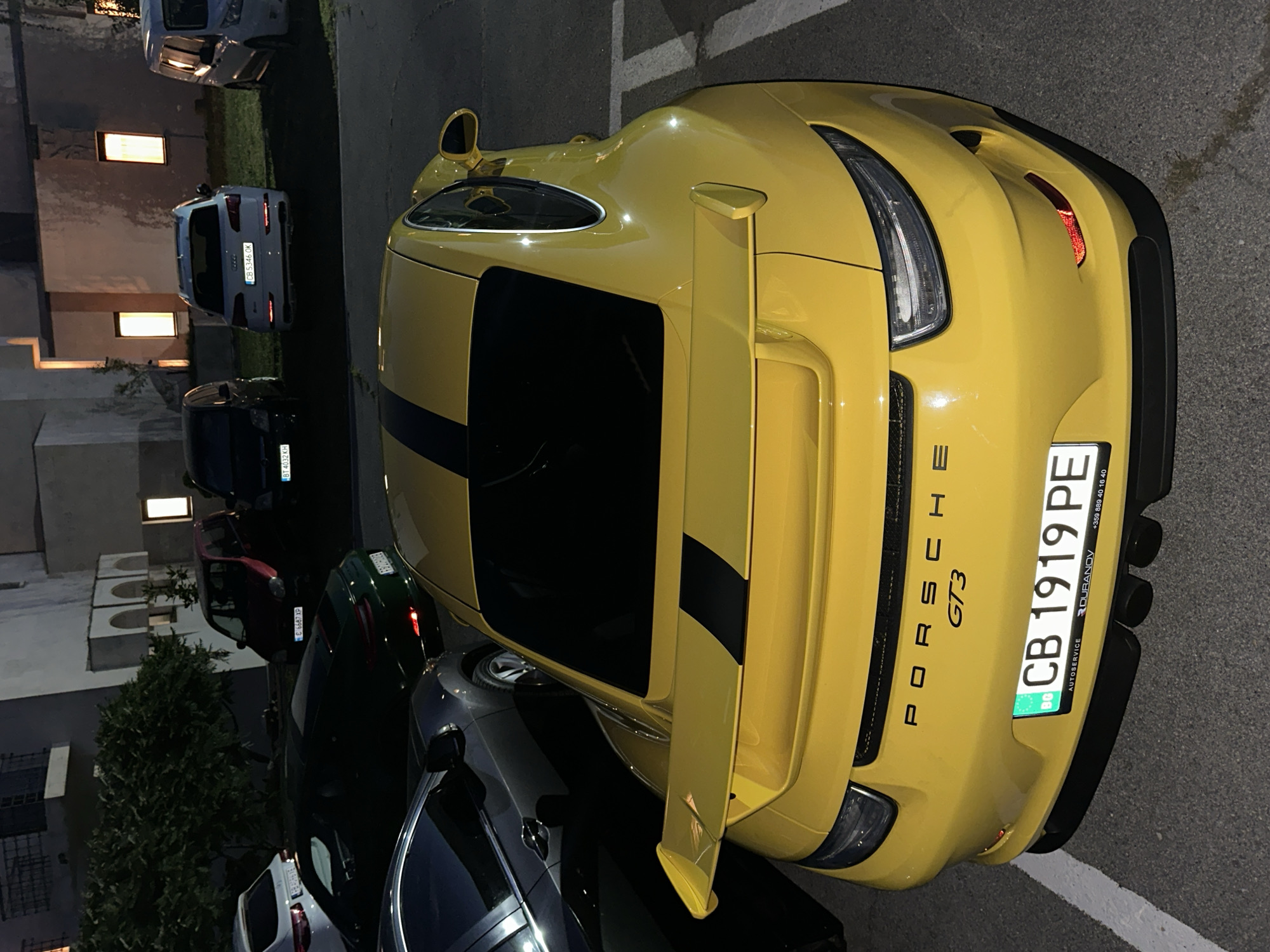 The image shows a yellow Porsche GT3 parked in an outdoor lot at night, likely in Arbanassi, Bulgaria. The car is the primary subject, with its rear-end prominently displayed. A black stripe runs down the center of the car's hood and roof. The car has a large rear spoiler and dual exhaust pipes. The license plate, "CB 1919 PE", is clearly visible and is from Bulgaria. In the background, other vehicles are parked, including a white SUV and a dark-colored van. Buildings with lit windows are visible behind the vehicles. The lighting suggests it is nighttime, and the ground appears to be asphalt. There are no people visible in the image.