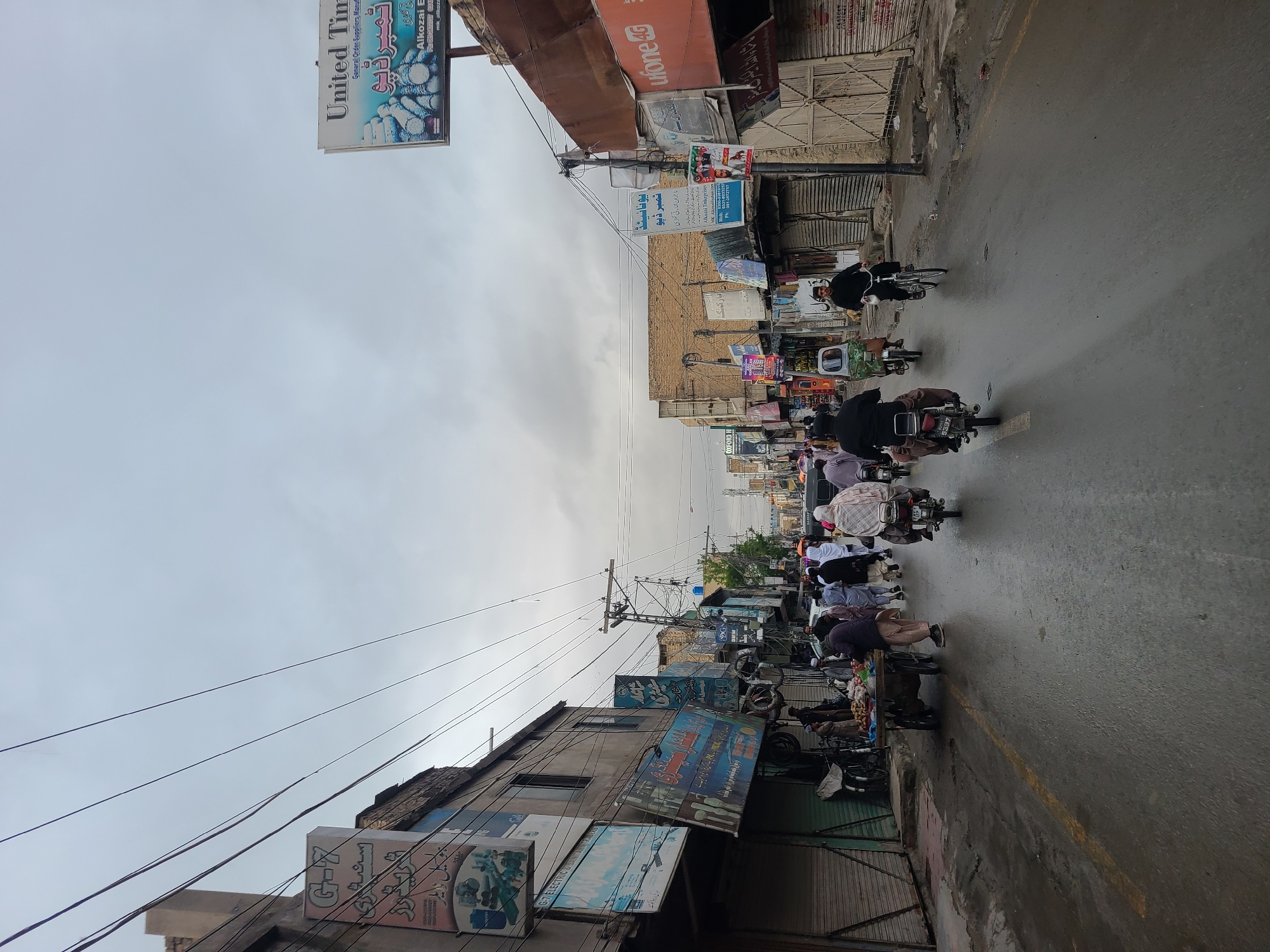 The image depicts a street scene, likely in Quetta City Tehsil, Pakistan, given the local signage. A row of people on motorcycles ride along a wet road, indicating possible recent rain. Storefronts line both sides of the street, with shop signs in Urdu and English. Overhead, power lines crisscross the sky. There are a few vendors on the sidewalks. The sky is overcast.