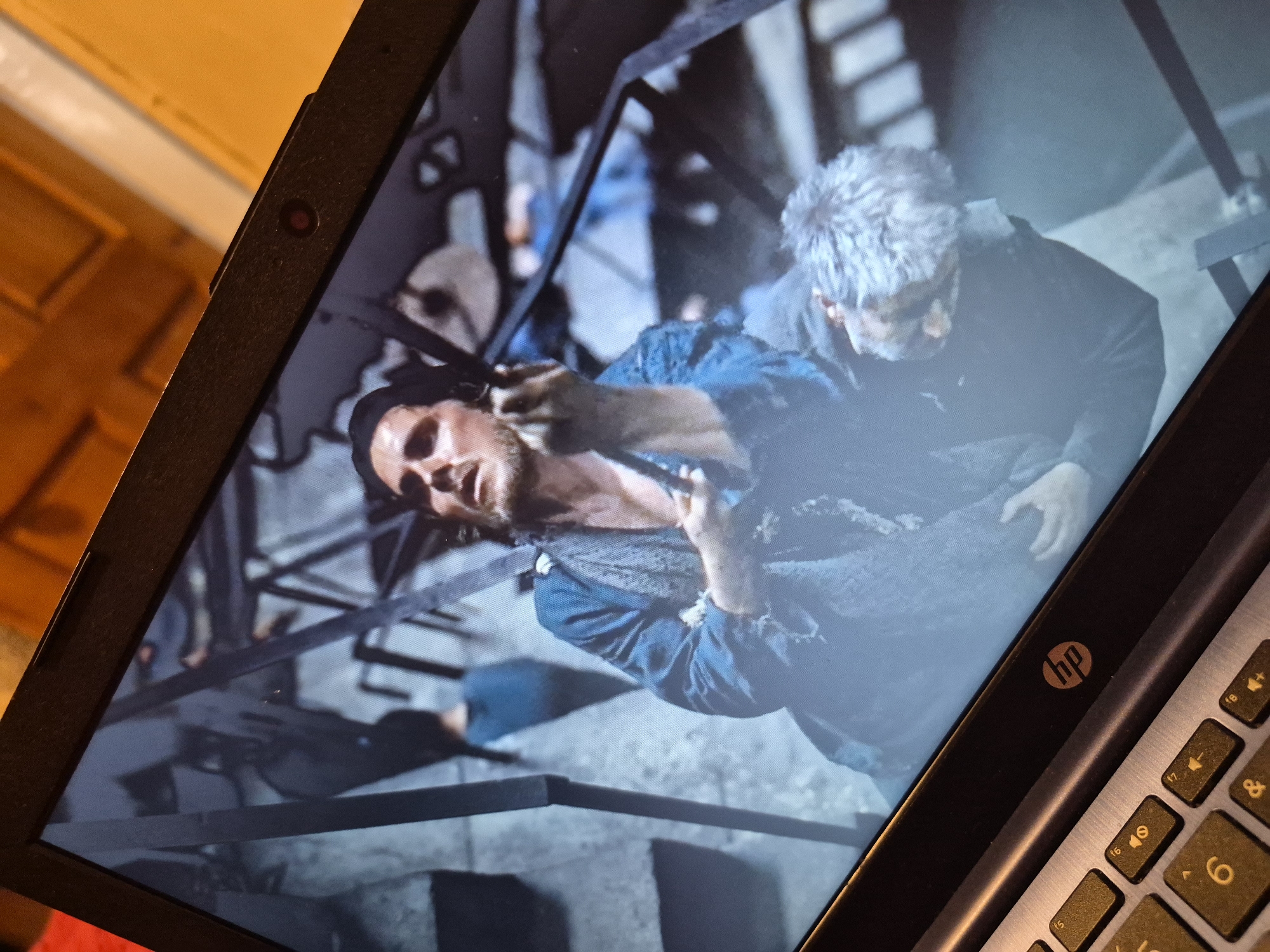 A laptop displays a scene from a film. The screen shows two men in what appears to be a distressed state. The man in the foreground, with dark hair and a beard, is wearing a torn blue garment and is looking upwards with his mouth slightly open. He is holding something in his hand. Behind him and to the right, another man with grey, messy hair is slumped against him, his face turned away. The background is blurry and suggests an industrial or urban setting with metal structures and possibly stone steps. The laptop itself is a silver HP model, partially visible in the foreground with a keyboard and the HP logo on the bezel. The image is taken from a slightly elevated angle, looking down at the laptop screen. The ambient lighting suggests an indoor environment, possibly a room. There is no visible indication of the location being in the city of Sandwell, United Kingdom.