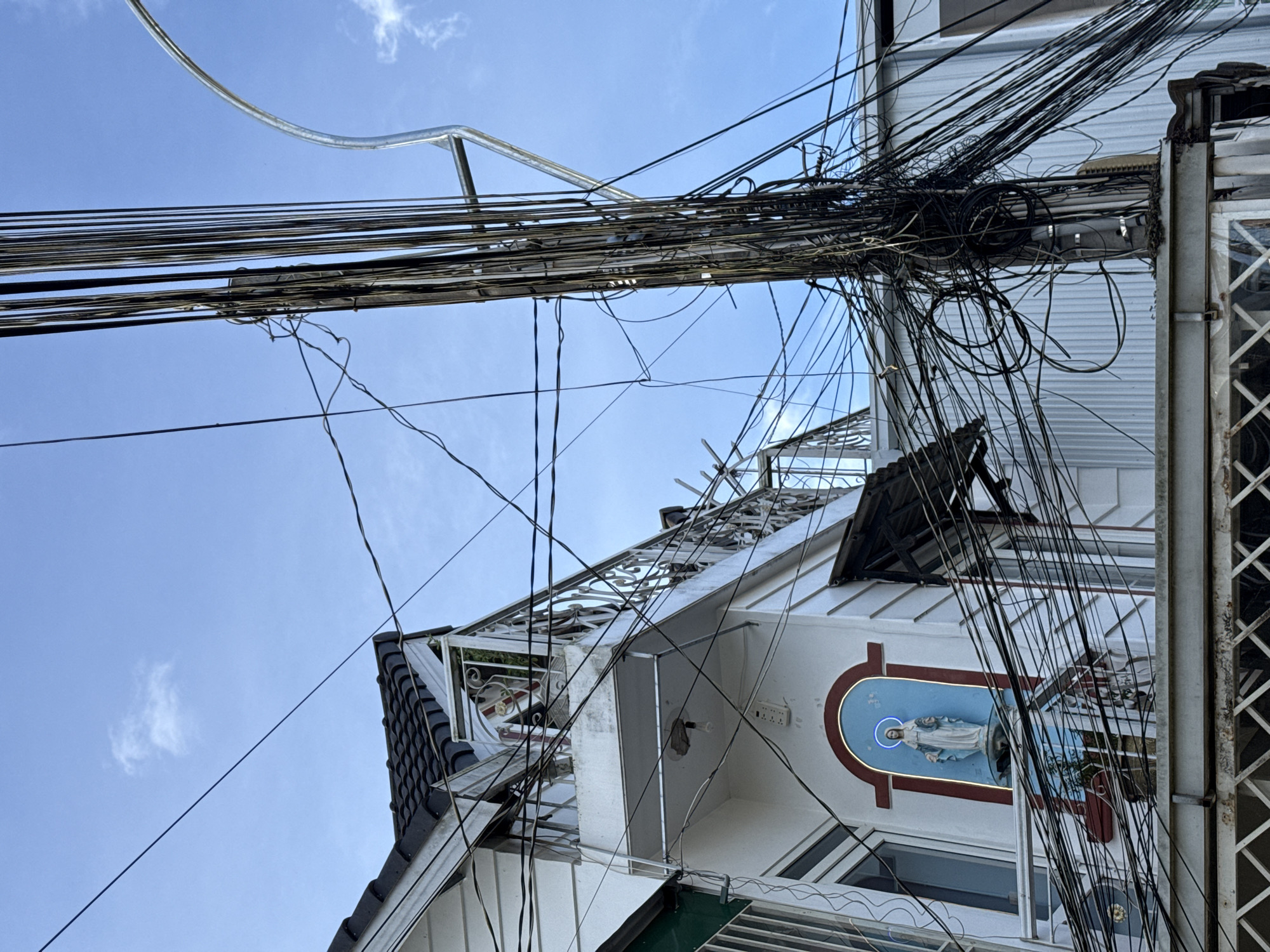 This image, taken in the city of Da Lat, Vietnam, depicts a chaotic tangle of electrical wires spilling from a utility pole onto a white building adorned with a religious statue. The building, with a black tiled roof and white siding, features a balcony with ornate metal railings. A statue of the Virgin Mary, illuminated by a blue neon halo, is positioned above an arched recess on the building's facade. The scene is dominated by a dense cluster of black wires, which crisscross the frame and obscure portions of the building and sky. A curved metal pole, possibly part of a street light, is visible on the right side of the image. The sky is a clear blue with a few wispy clouds. The overall impression is one of urban infrastructure in disarray, with the religious iconography providing a stark contrast to the surrounding electrical clutter.