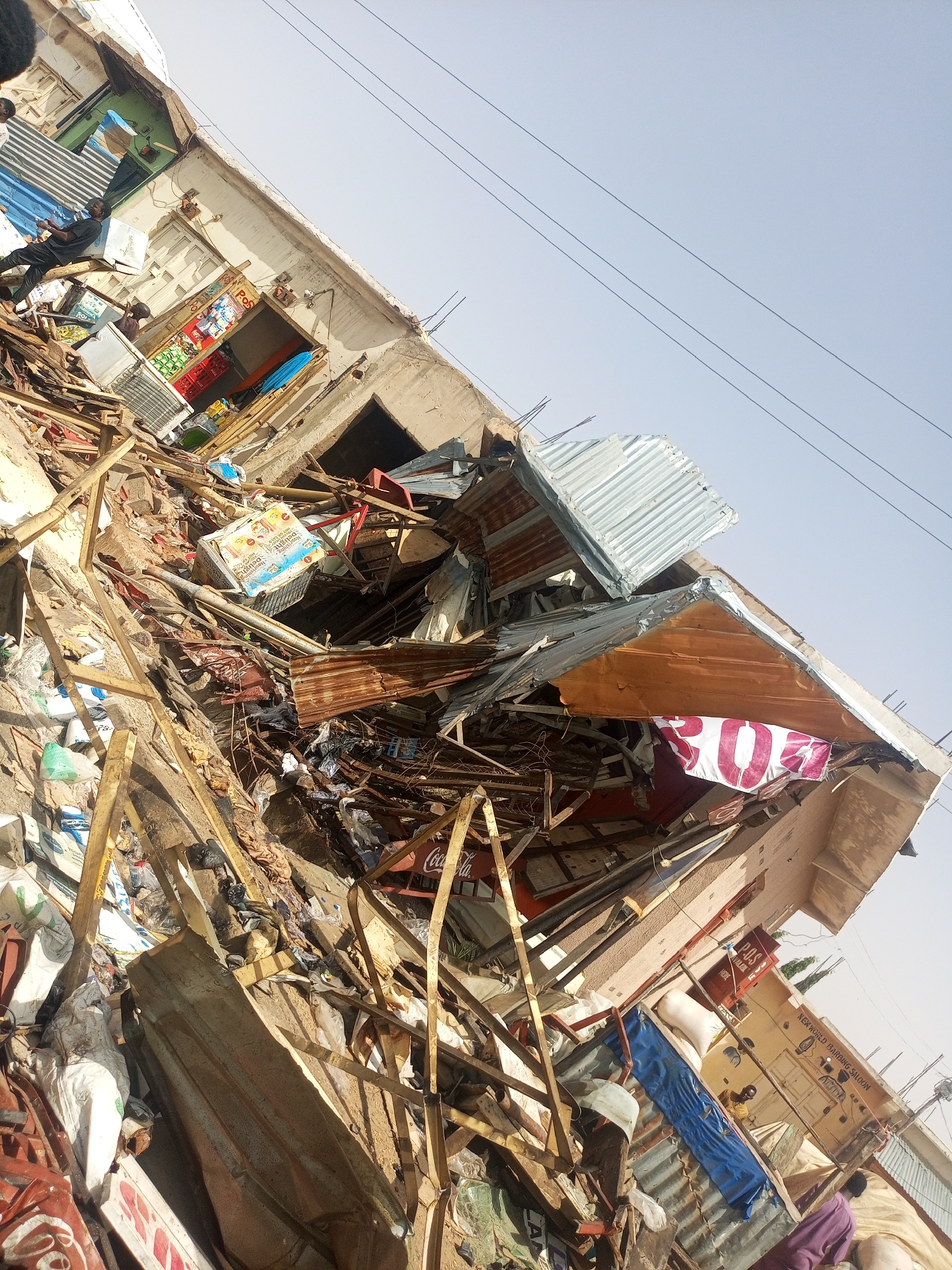 The image depicts a scene of significant destruction, likely in Gombe, Nigeria. A building has partially collapsed, scattering debris consisting of metal sheets, wooden beams, and various household items. Among the debris are commercial elements such as "Coca-Cola" and "POS" (Point of Sale) signage. Several individuals appear to be navigating or assessing the scene, their presence suggesting a post-disaster environment. The sky is clear, indicating daylight conditions. The environment shows signs of a commercial area.