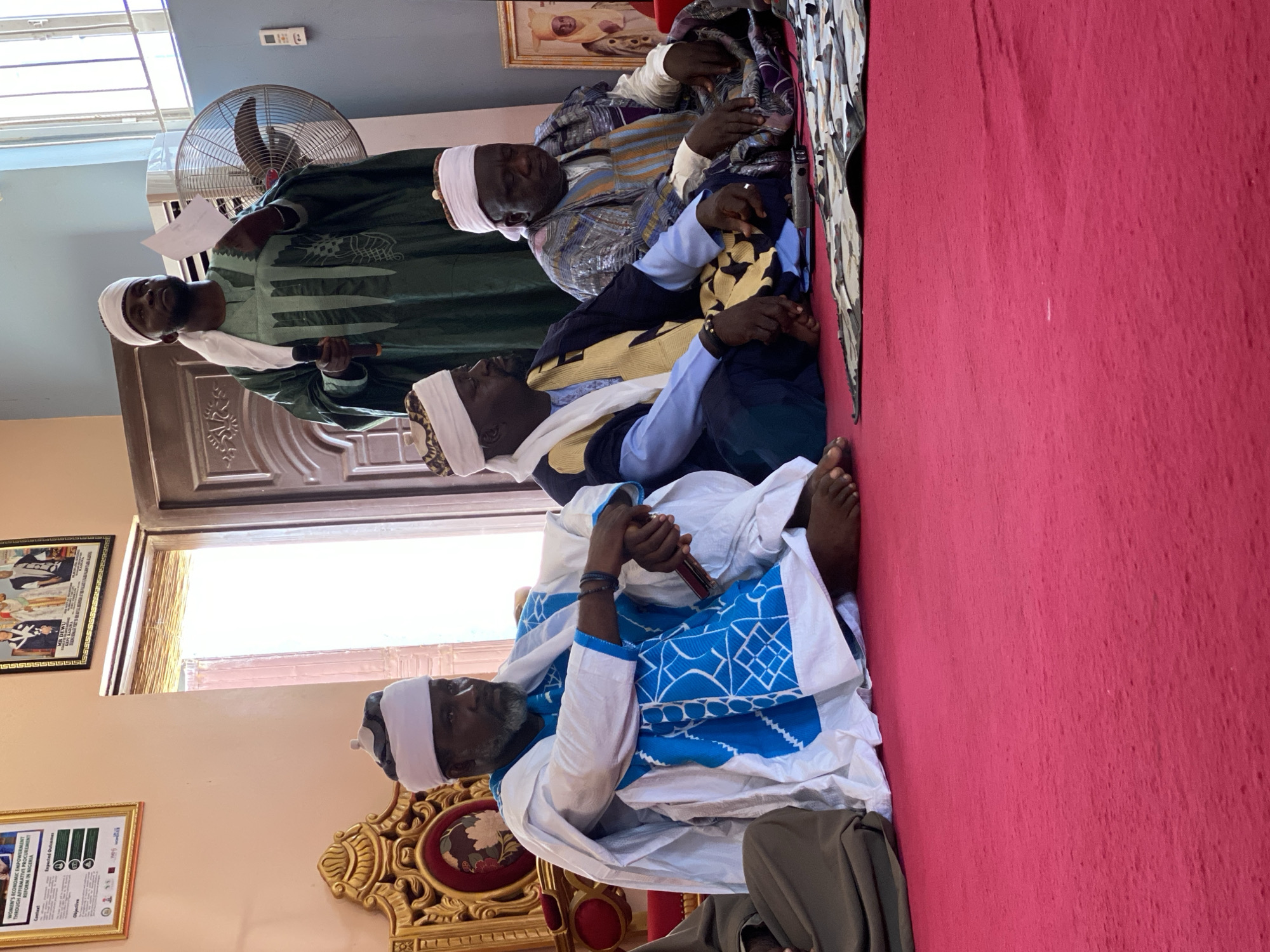 Here is a detailed description of the image:
**People and Interactions:** The image shows five men, all dark-skinned, likely of African descent, gathered in a room. Four of them are seated, possibly on the floor, while one man stands. All men are wearing traditional-looking robes and headwear. The man standing is holding a piece of paper in one hand and a microphone in the other, suggesting he may be speaking or giving a presentation.
**Objects:** There is a large fan visible in the background. A red carpet covers the floor area where the men are gathered. A picture frame is mounted on the wall, next to a window. The bottom of a poster can be seen, containing an image of what appears to be some type of document or flyer.
**Environment:** The setting appears to be indoors, likely within a home or community building. The presence of framed pictures and decorated furnishings suggests a more formal or gathering space.
**Location:** The image's OCR references "REFORM IN NIGERIA" which would indicate this photo has been taken in the city of Sabon Tasha, Nigeria.