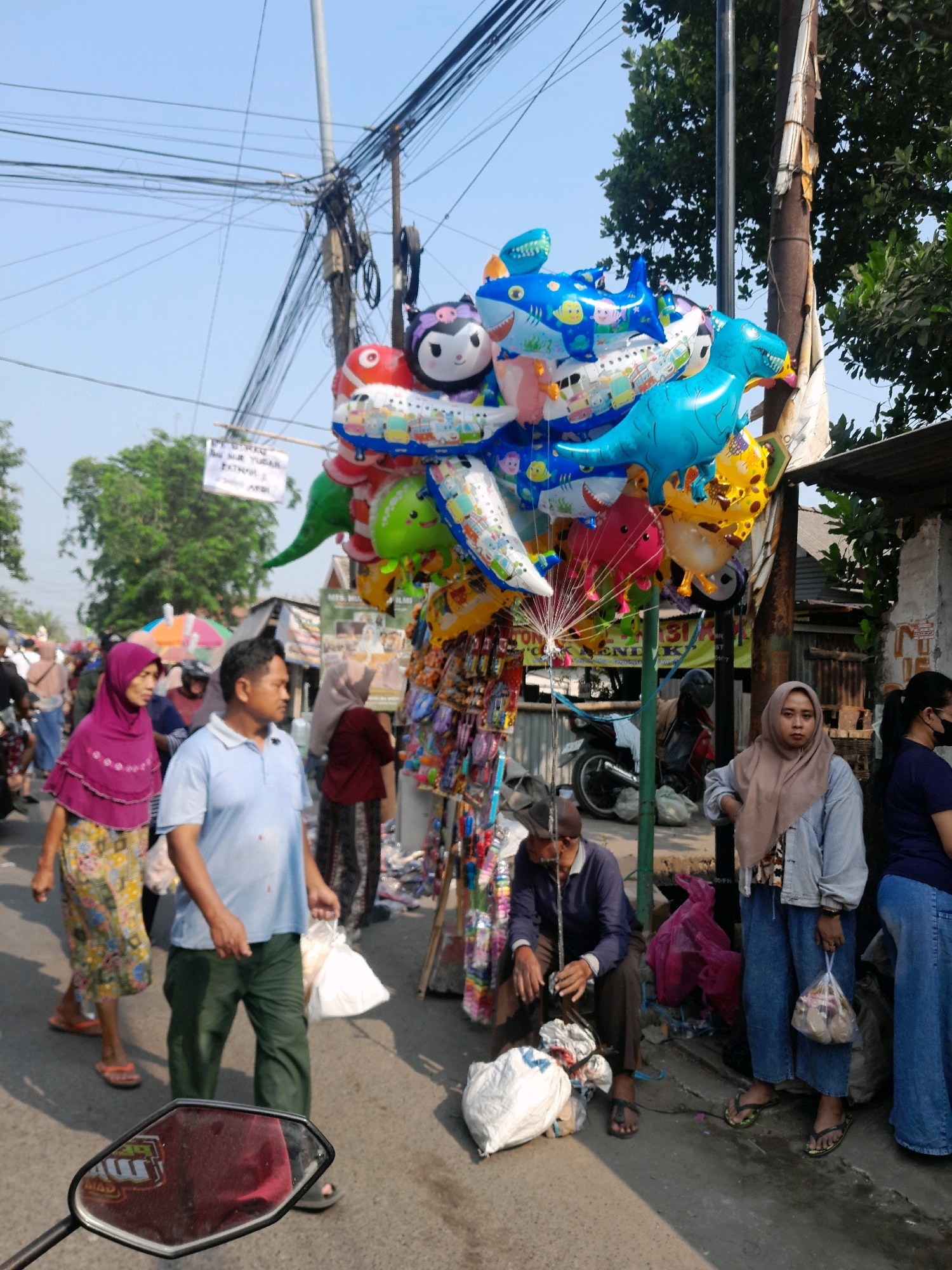 The image depicts an outdoor market scene in Jogosatru, Indonesia, with a focus on a vendor selling balloons. In the foreground, the reflection of a red object, possibly a vehicle or a bag, is visible in a mirror. A man wearing a light blue polo shirt and green pants is walking towards the right, carrying a white plastic bag. Beside him, a woman in a purple hijab and a patterned skirt is also walking.
Behind them, a large cluster of colorful balloons, including animal shapes like sharks and dinosaurs, as well as character balloons, are suspended from a pole. A vendor stall with various items, likely snacks or trinkets, is displayed next to the balloons. Several people are browsing or walking through the market. A man in a dark blue long-sleeved shirt and brown pants is sitting down, holding a white bag. Another woman in a light brown hijab and a gray jacket is standing and holding a bag. The background shows a street with trees, utility poles, and buildings, suggesting a bustling urban environment. The sky is clear and blue.