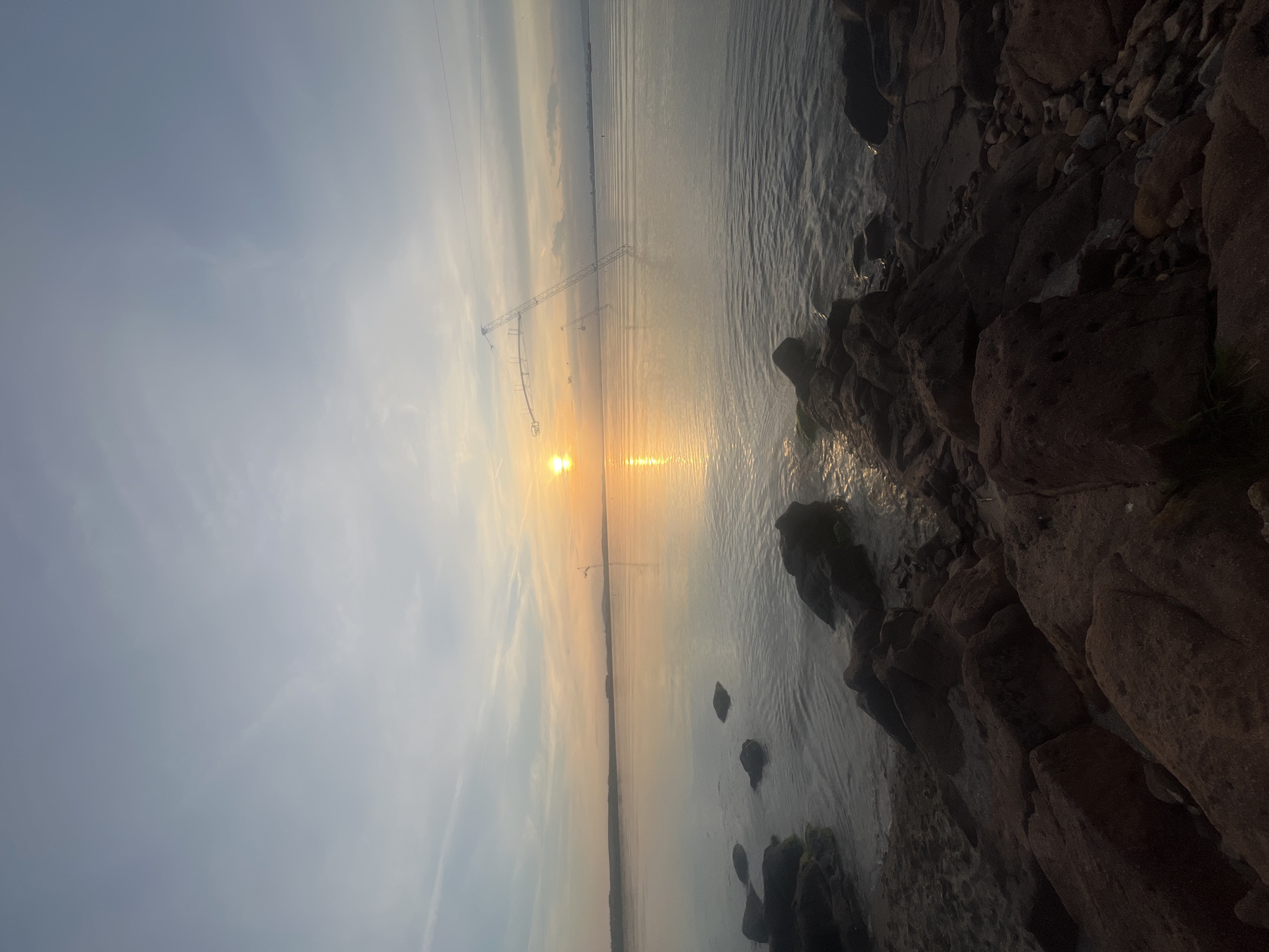 The image shows a body of water meeting a rocky shoreline, with the sun reflecting off the water's surface. In the distance, power lines stretch across the water. The sky is overcast with varying shades of light gray and yellow. The location is likely Burgas, Bulgaria, given the presence of the sea and power lines characteristic of coastal areas. There are no people visible in the image. The primary subjects are the water, rocks, sky, sun, and power lines. The interaction occurs between the sunlight and water creating a reflection. The environment appears to be natural with rocky terrain meeting the sea.