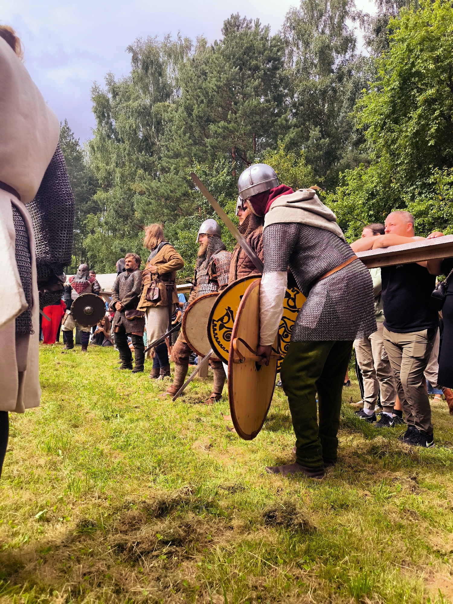 In Tērvete, Latvia, a group of people are gathered on a grassy field, some dressed in medieval armor and tunics, engaged in a reenactment. The foreground is dominated by a person wearing a chainmail tunic, helmet, and holding a wooden shield. Behind them, other individuals in similar historical attire stand with swords and shields. In the background, trees form a dense natural backdrop, with a clear sky overhead. Spectators, some leaning on a wooden barrier, observe the reenactment. The overall atmosphere suggests a historical festival or demonstration.