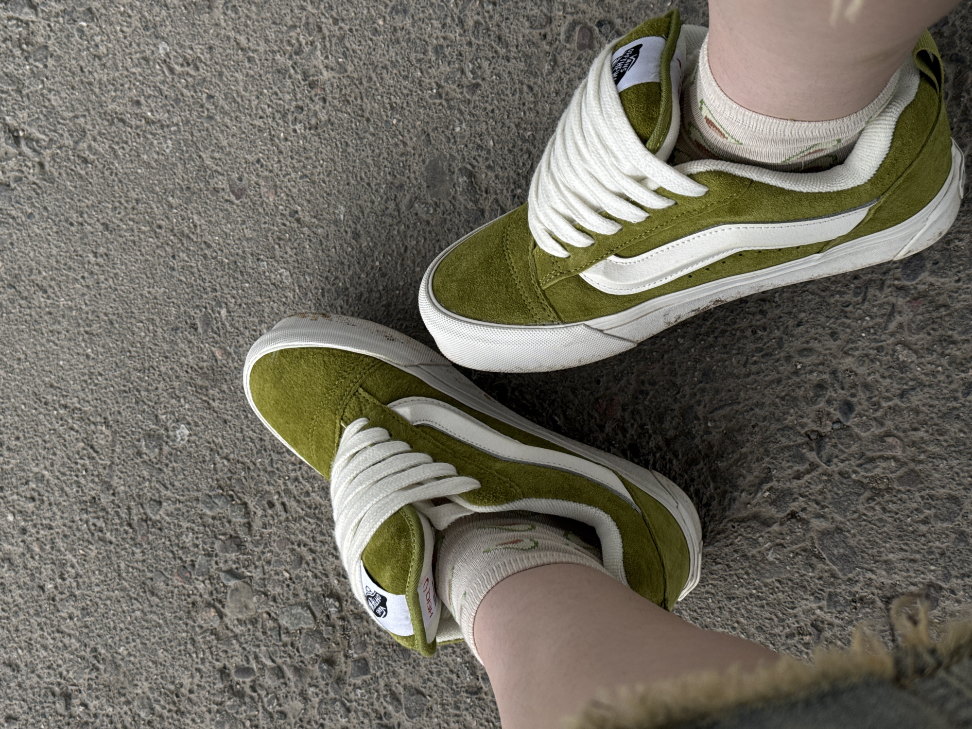 A top-down, medium shot shows a pair of olive-green Vans sneakers with white laces and a white side stripe, worn with white socks with a floral pattern. The shoes are on a person's feet, and the feet are resting on a rough, gray asphalt surface. The ground is textured with small stones and pebbles. There is no indication of the specific location within the city, nor any signs of human interaction or activity beyond the person wearing the shoes.