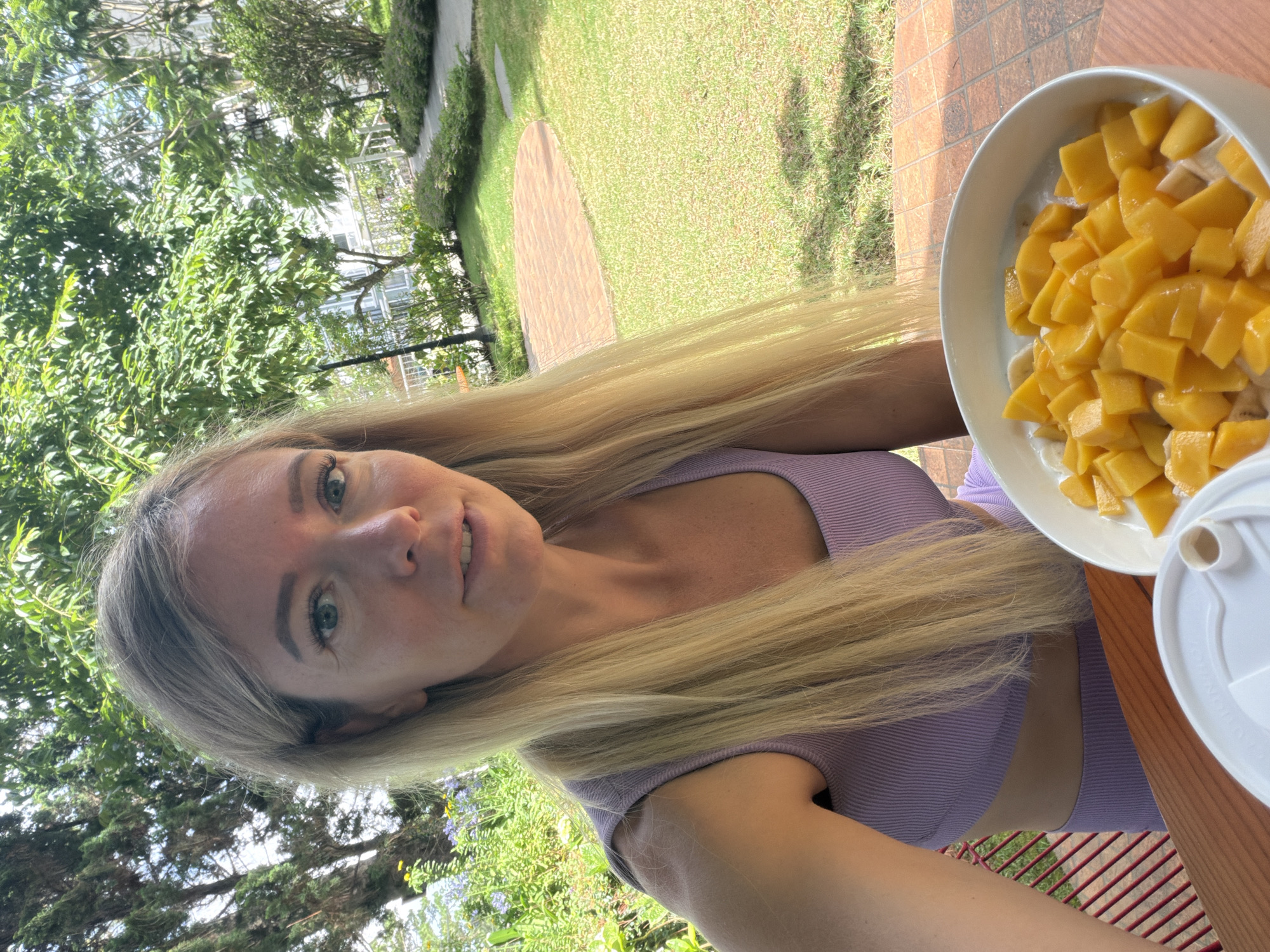 A blonde woman is seated at a wooden table outdoors, with a bowl of chopped mango and what appears to be yogurt or ice cream in front of her. She is wearing a lavender ribbed crop top and shorts. Behind her, a lush green garden with trees and a pathway is visible, suggesting a tropical or subtropical setting. The location is identified as Xuân Trường, Đà Lạt, Vietnam.