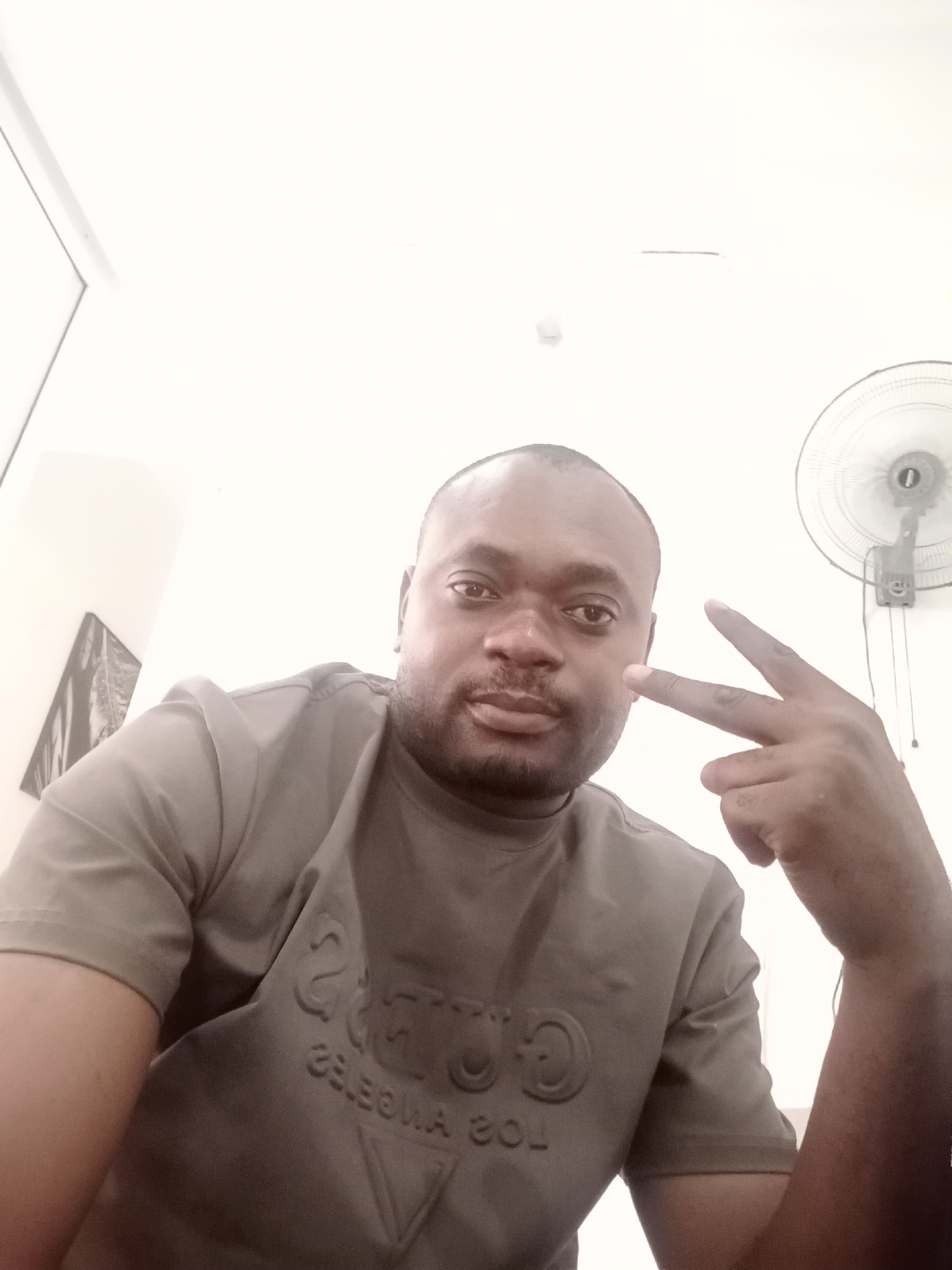 A man is positioned indoors with a standing fan visible in the background. He is making a peace sign with his right hand. He is wearing a short-sleeved olive green shirt with the word "GUESS" and "LOS ANGELES" in raised lettering. The setting appears to be Gwarinpa, Nigeria. There is artwork visible on the wall behind him.