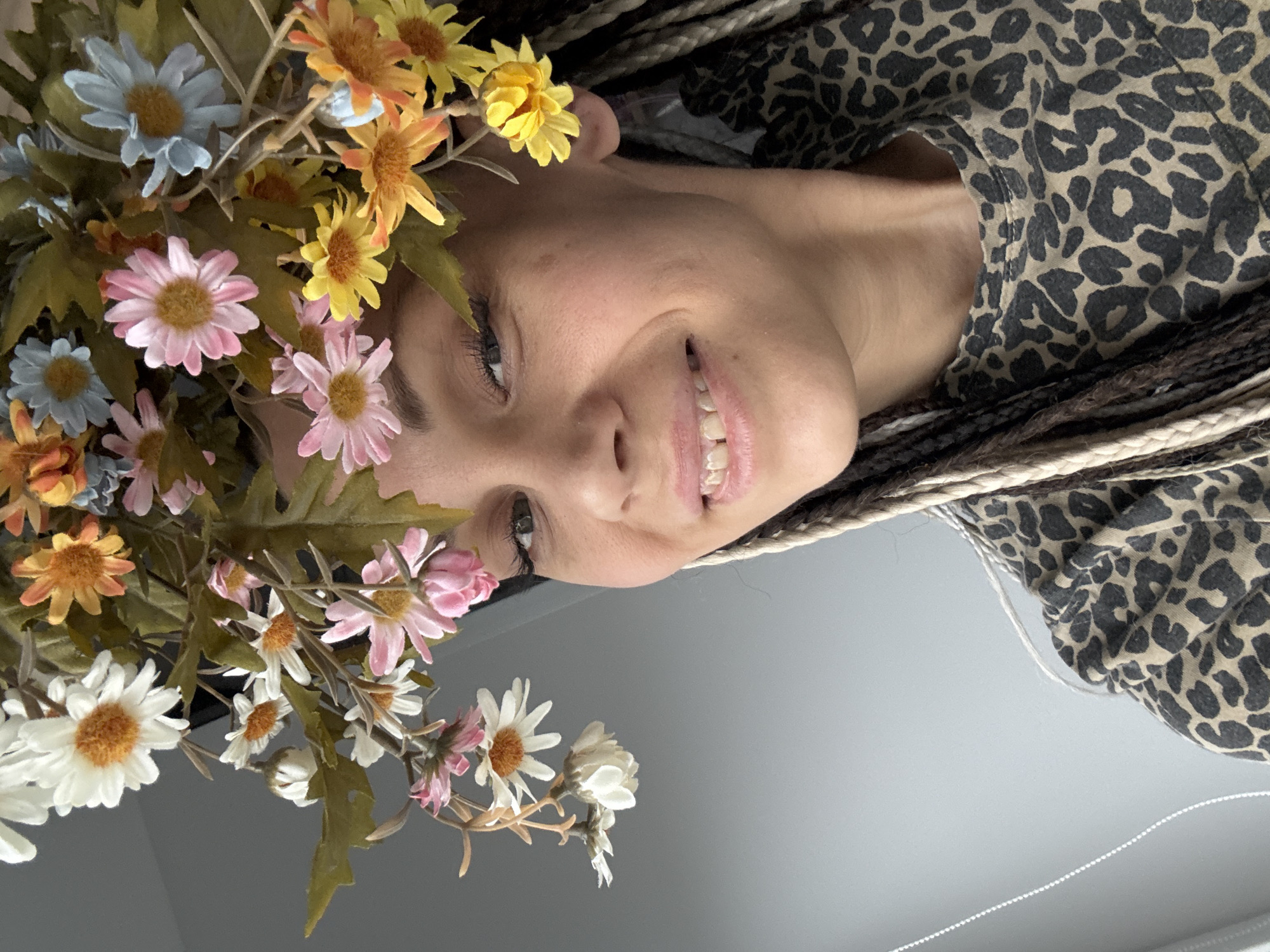 A person with brown and white braids is shown smiling upside down. The person is wearing a leopard print top. There are several flowers in various colors, including white, pink, orange, and blue, near the person's head. The background is a smooth gray surface. It is unclear whether the image was taken in Rusanovo, Russia.