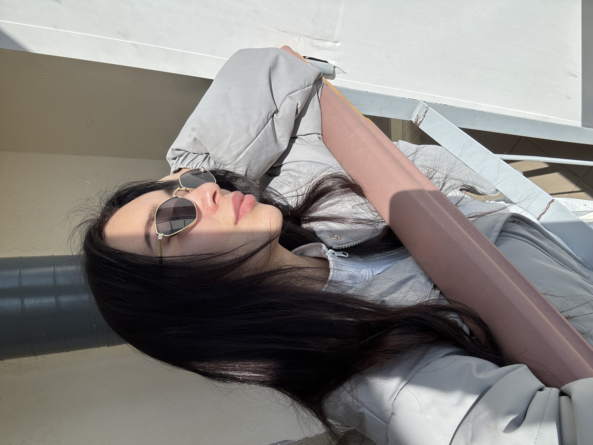 A woman lies on her back, wearing sunglasses and a light gray jacket, with dark hair spread around her. She is positioned on what appears to be an outdoor structure with white railings and a beige background, suggesting a balcony or elevated area. The setting could potentially be Chaikovsky, Russia.