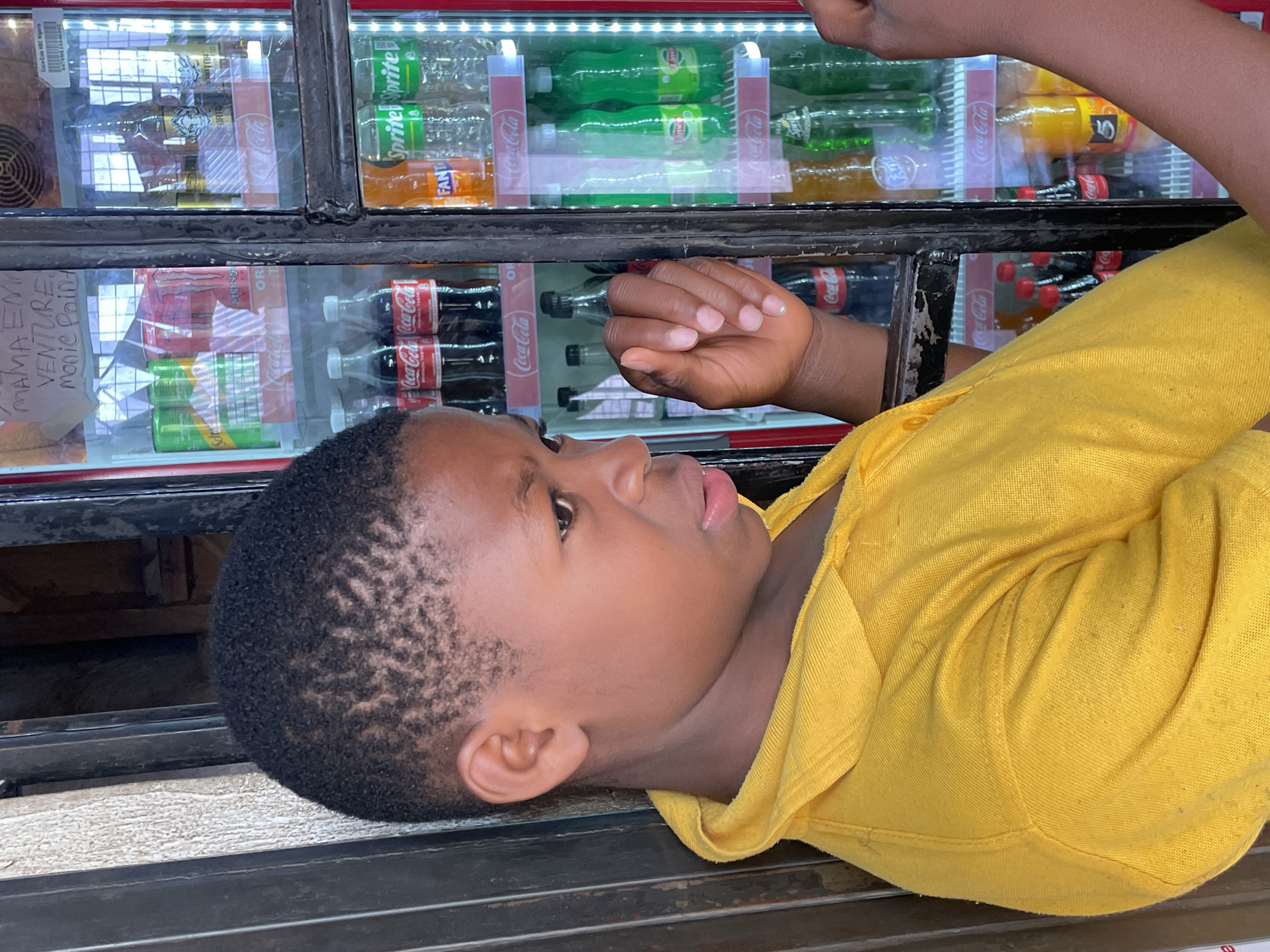 A young boy with short, dark hair wearing a yellow shirt lies on a metallic surface, positioned near a refrigerator displaying various bottled drinks, including Coca-Cola, Sprite, and Fanta. A handwritten sign reading "MAMA EM VENTURE Monie Paint" is affixed to the refrigerator. The boy's eyes are open, and his hand is near the cooler glass. The location is likely a retail or store setting in Jos, Nigeria.