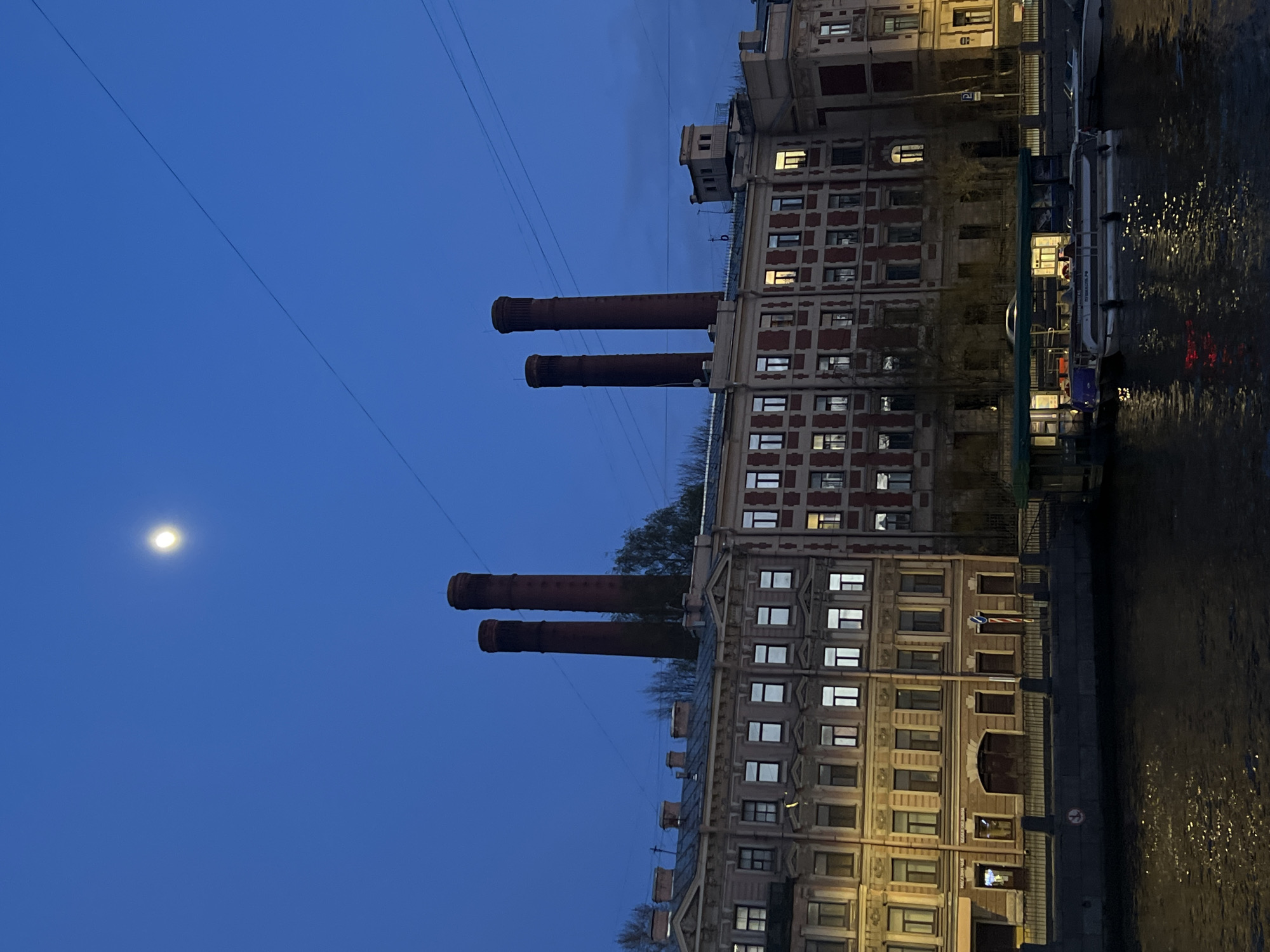 This media file captures an urban waterfront scene at twilight or night in Saint Petersburg, Russia. A deep blue sky dominates the upper portion of the image, where a bright, full or near-full moon is visible in the upper left. Several thin, dark overhead wires stretch across the sky.
On the right, a large, multi-story building with classical architectural features lines the bank of a canal. Its facade combines light-colored and reddish-brown sections, and many of its numerous windows are illuminated with warm yellow light. From behind or on the roof of this building, two prominent, tall, reddish-brown industrial chimneys rise into the sky. A smaller, simpler structure is also visible on the rooftop further along the building.
The canal occupies the lower part of the frame, its dark water reflecting the building lights and showing subtle ripples. A passenger boat or ferry, illuminated from within, is docked or cruising on the canal near the building, with another vessel partially visible.
An embankment runs alongside the canal in front of the building. On the building's facade, faintly visible Cyrillic text "МАГАЗИН ЧАЯ" (Tea Shop) can be seen on a sign, along with a blue 'P' (parking) sign. A round red and white regulatory sign is also present on the embankment. Bare tree branches are discernible behind the chimneys and buildings, suggesting a cool season. The scene conveys a calm and serene evening atmosphere in the city.