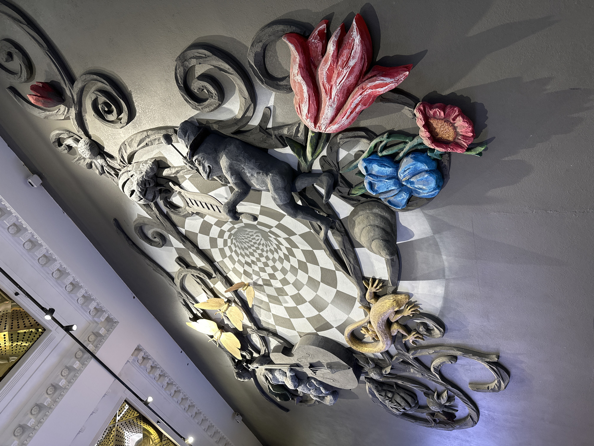 The image displays a highly detailed, fantastical high-relief sculptural installation adorning a ceiling or upper wall, likely in an indoor public space in Yekaterinburg, Russia. The artwork combines whimsical and surreal elements against a dark grey background, centered around a swirling checkerboard vortex design.
Prominently featured is a grey, anthropomorphic figure, possibly a monkey or similar creature, wearing a hat and appearing to play a keyboard-like instrument. Nearby, a large, textured red tulip draws the eye, accompanied by smaller blue bell-shaped flowers and a pink bloom. Further down, a golden lizard or salamander is intricately sculpted, coiling among the relief's foliage. Several golden butterflies or moths are depicted near a stylized stringed instrument, resembling a lute or mandolin. The entire composition is framed by elaborate, dark grey swirling patterns that enhance its three-dimensional quality and sense of movement.
The sculpture is illuminated by artificial lights, casting dramatic shadows that accentuate its depth and texture. The vibrant colors of the flowers and golden creatures contrast effectively with the predominantly grey tones of the figures and background. On the left side of the frame, glimpses of the surrounding architecture reveal ornate white moldings, modern track lighting, and golden perforated panels, suggesting an opulent or artistically designed interior. No visible text is present in the image.